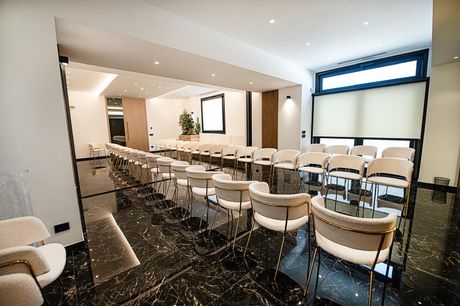 Sala conferenze moderna con file di sedie bianche, lunghi tavoli e uno schermo di proiezione.