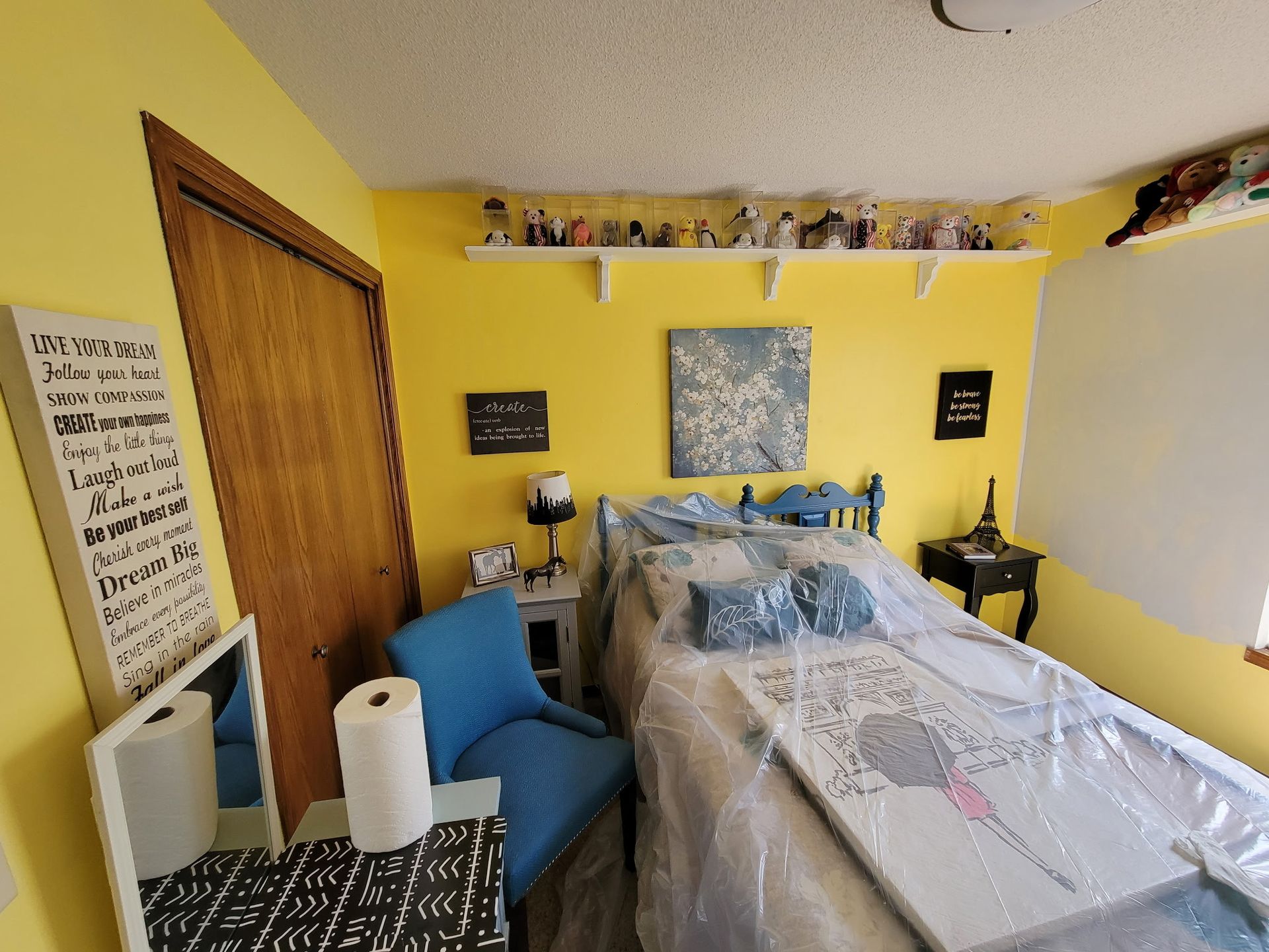 Yellow bedroom with a bed covered in plastic, a blue chair, and various decorations.
