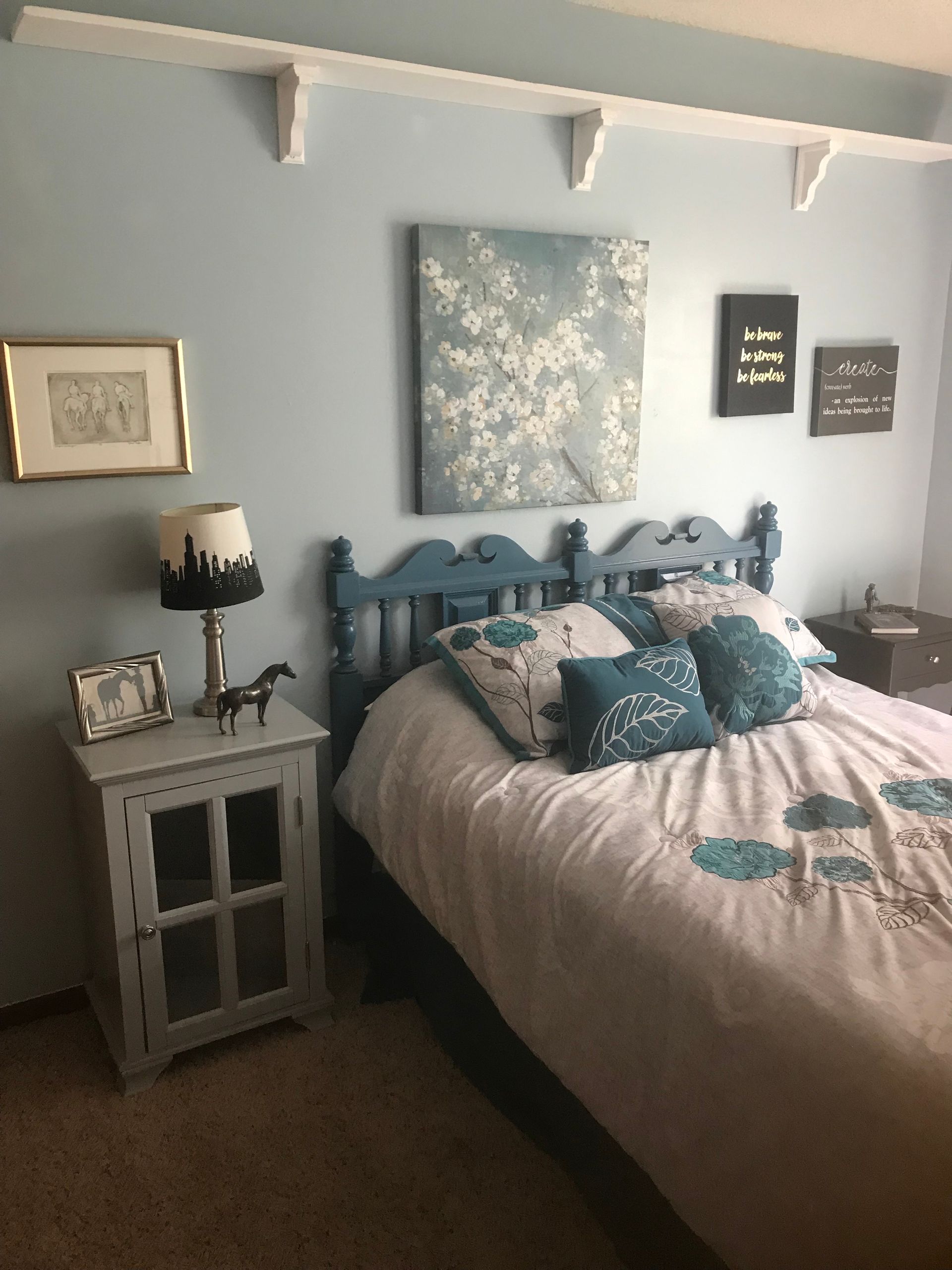 Bedroom with blue walls, bed with teal accents, nightstand with lamp, and artwork.