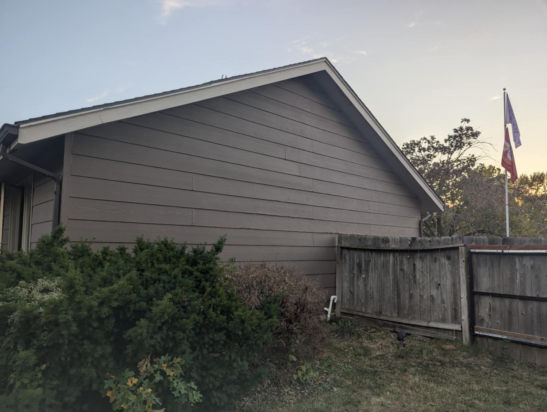 Brown house with tan trim, wooden fence, bushes, and a flag on a pole.