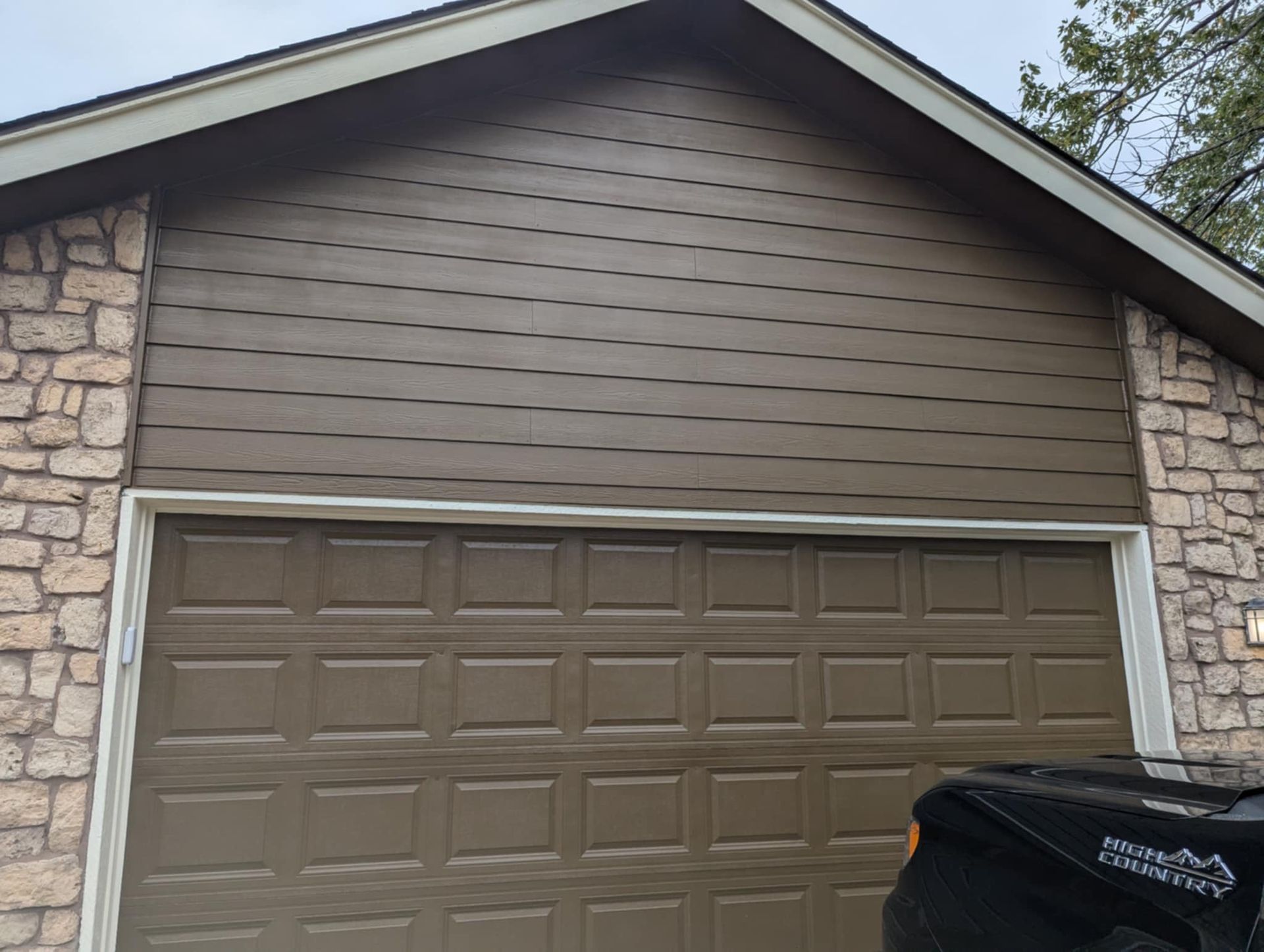 Brown garage door with matching siding, set against stone walls, under a cloudy sky.