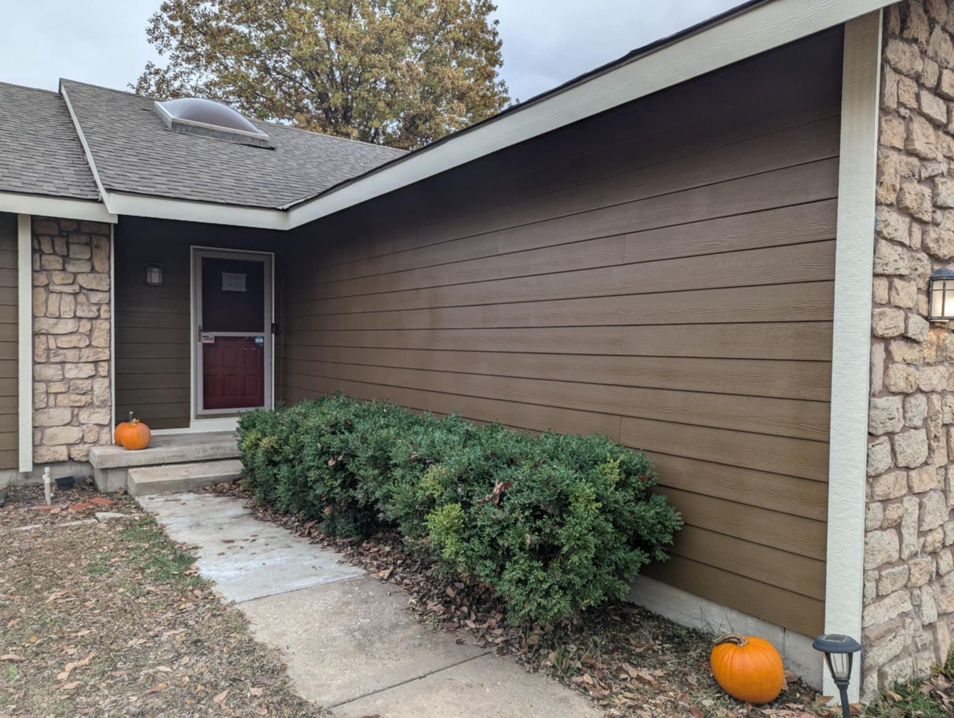 Brown house with stone accents, small porch, green bush, and pumpkins on the path.