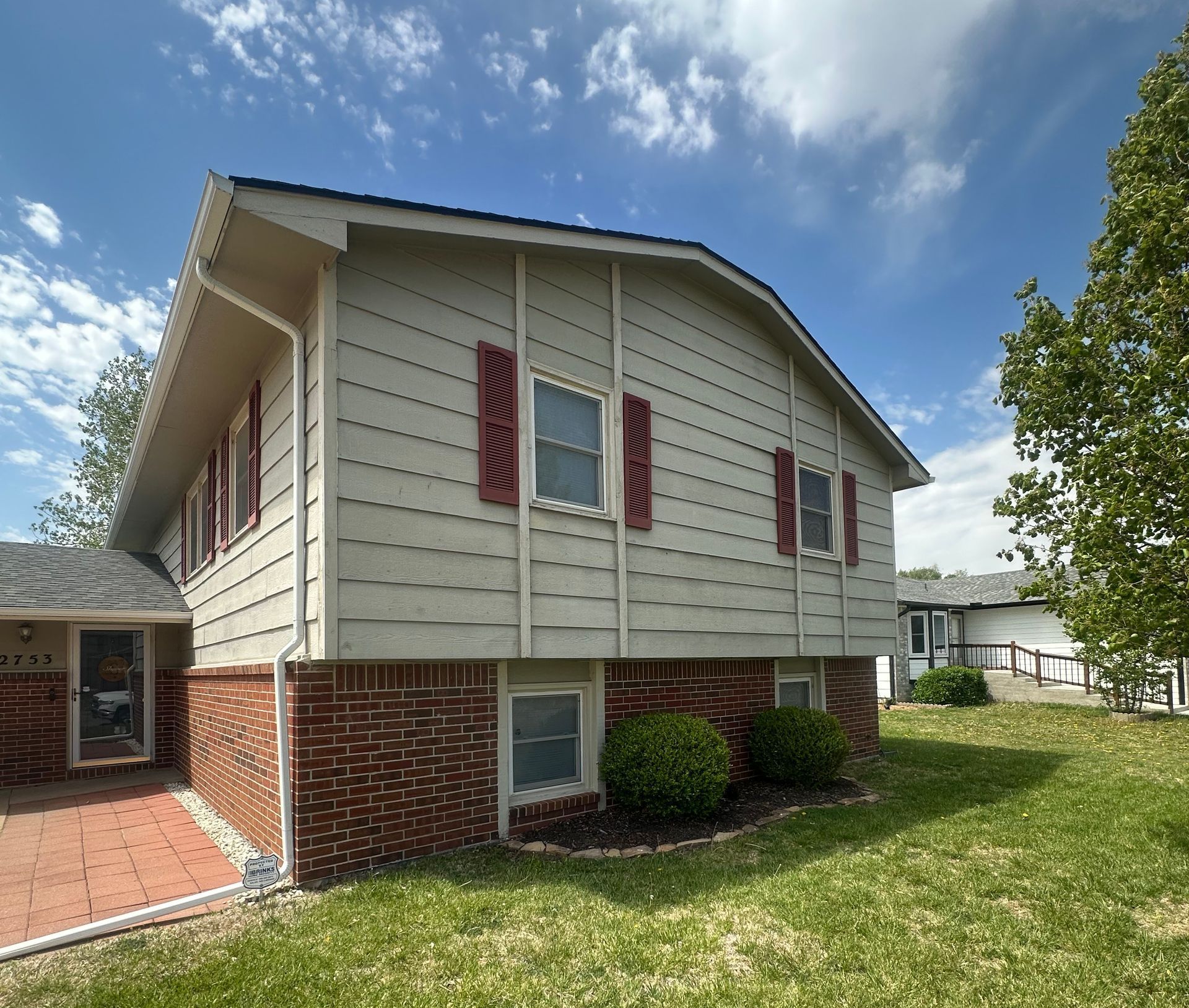 Side view of a two-story house with red shutters and a brick base, under a blue sky with some clouds.