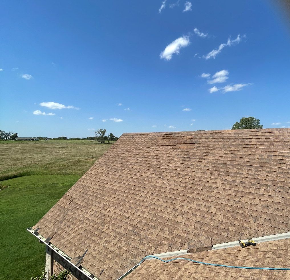 Brown shingled roof against a blue sky with sparse clouds, overlooking a grassy field.