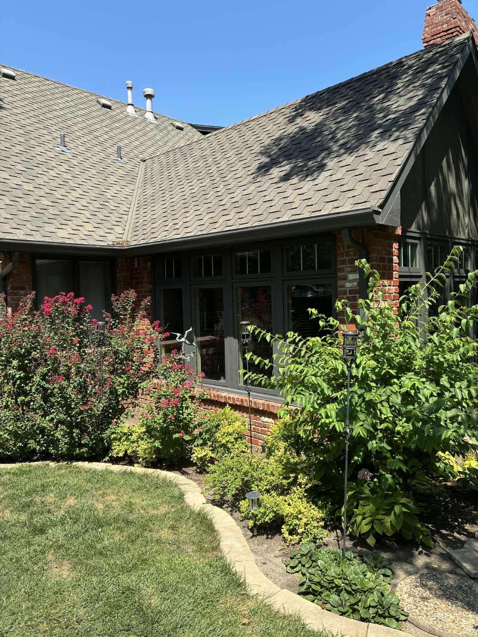 Lush shrubs and a green lawn frame a brick house with a grey roof under a blue sky.