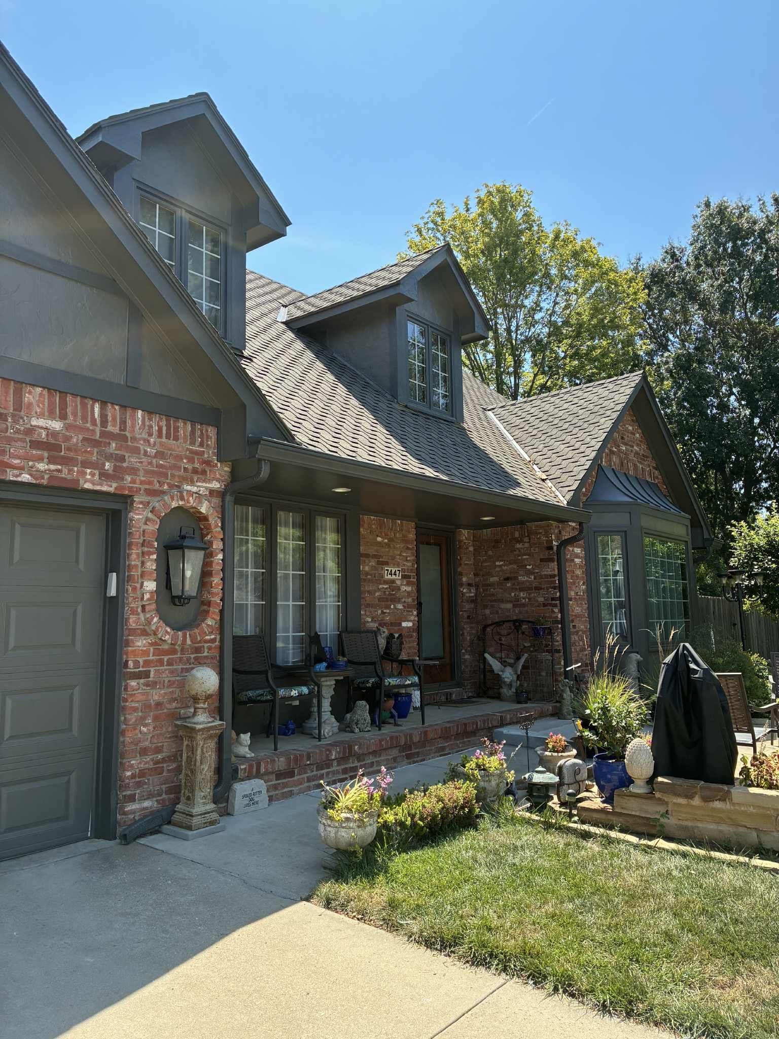 Brick house with gray roof, dormers, and garage. A front porch with chairs, flower beds, and lawn.