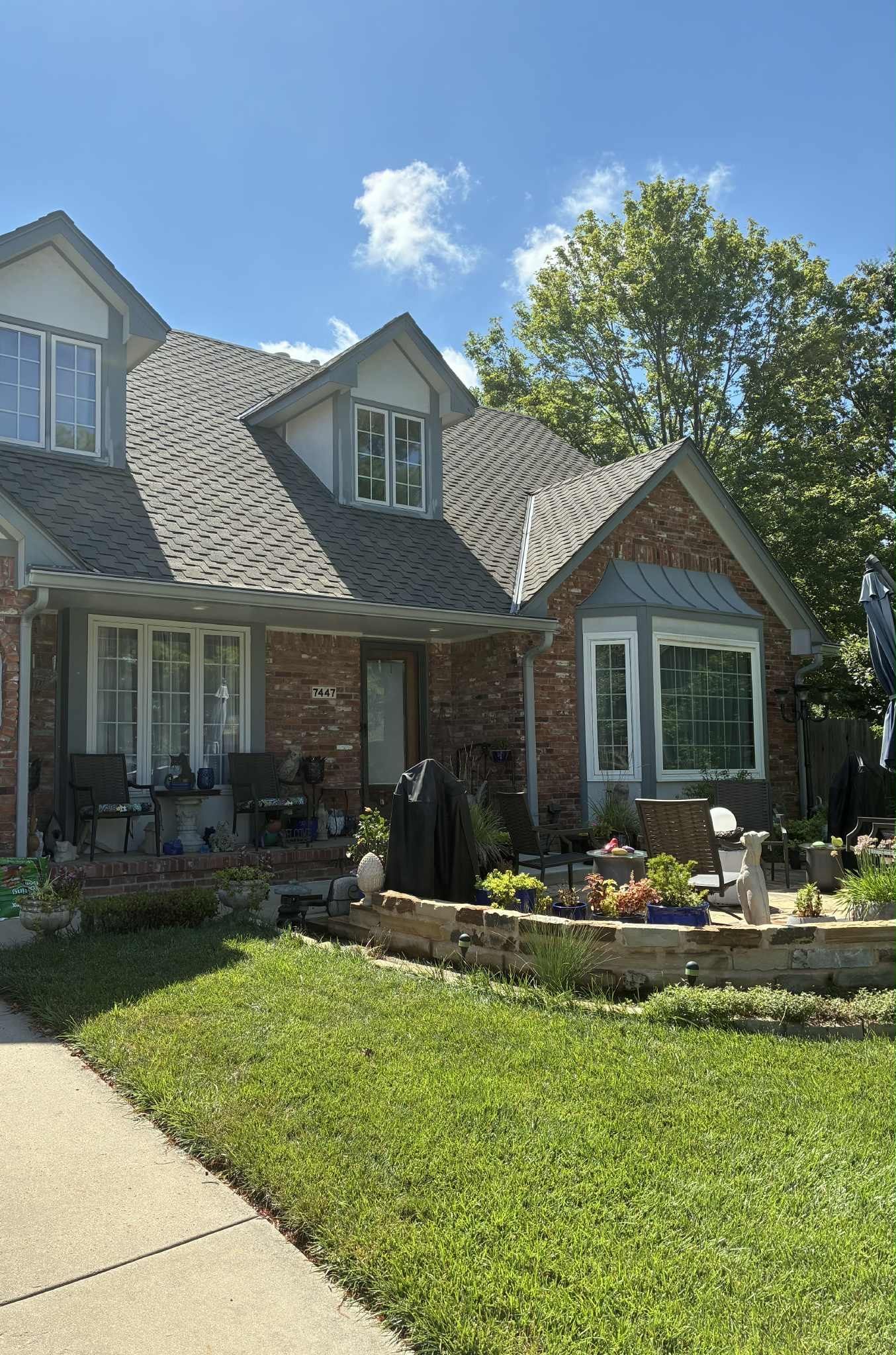 Brick house with a gray roof, dormers, and a lush front yard with a sidewalk.