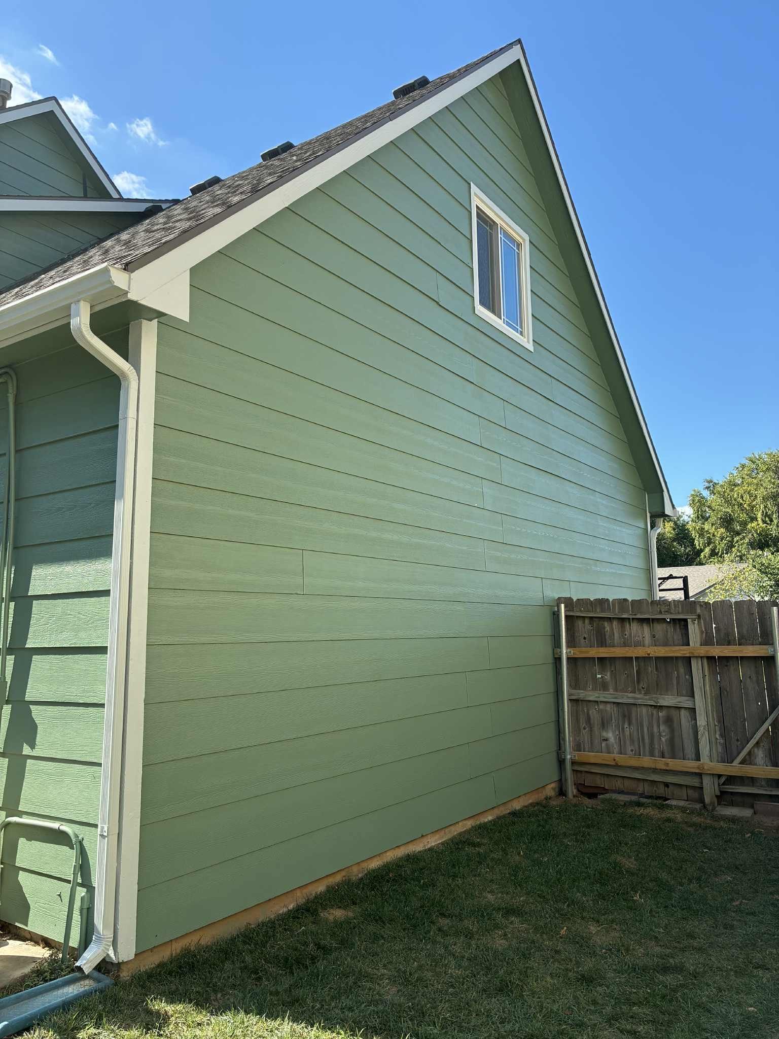 Green house siding with white trim, a small window, and wooden fence against a blue sky.