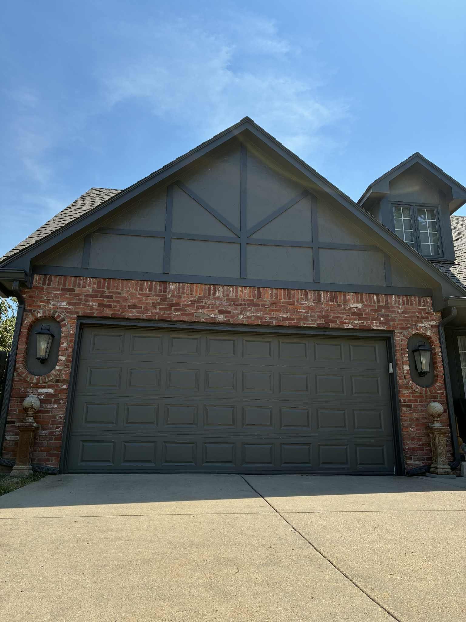 A garage door with gray paint, brick facade, and a dark gray gable roof under a blue sky.