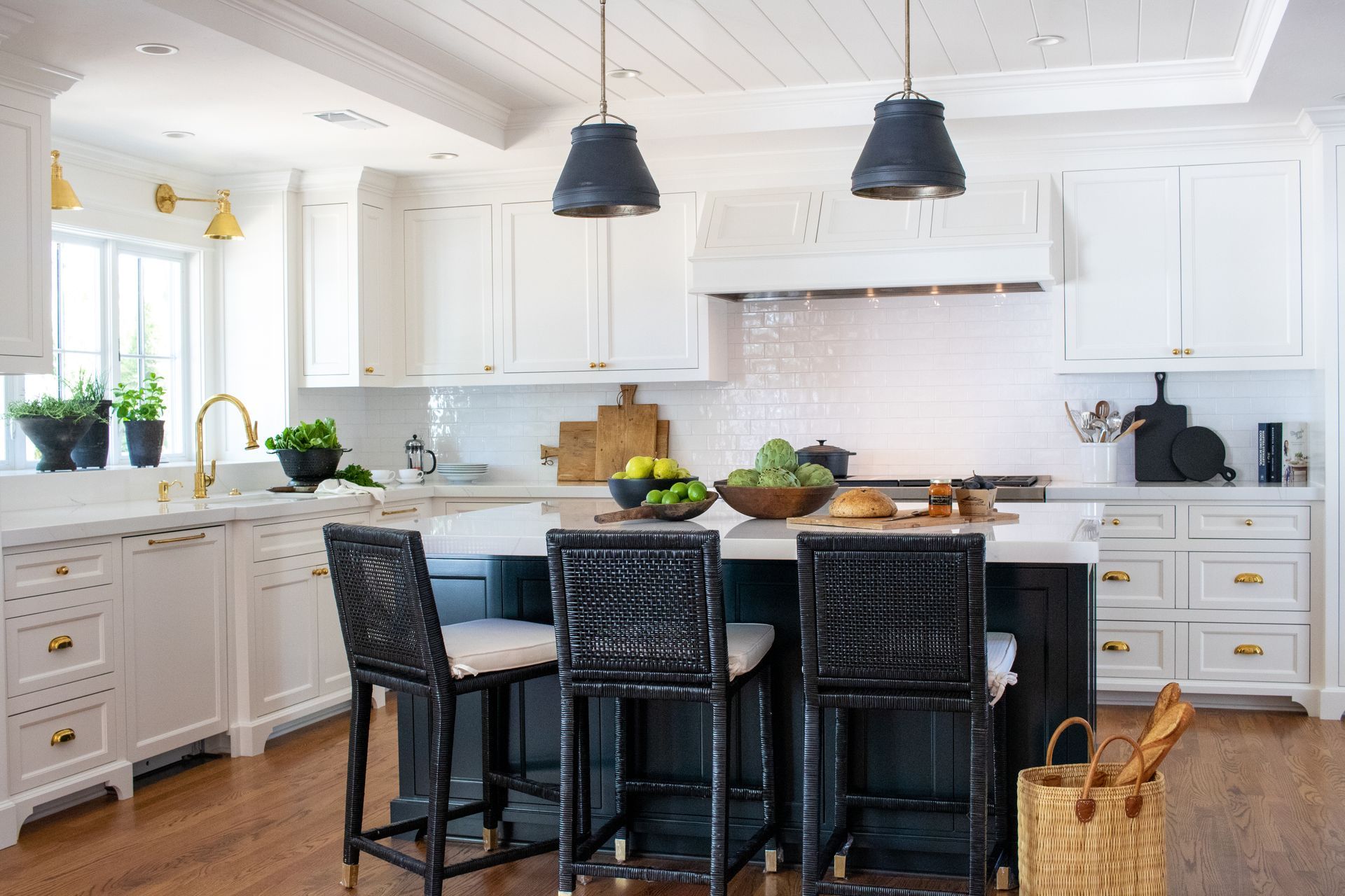 White kitchen with dark island and black wicker bar stools. Gold hardware and pendant lights.