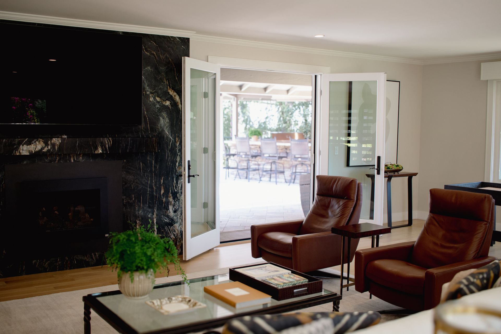 Living room with brown leather chairs, fireplace, and open doors to a patio with tables and chairs.