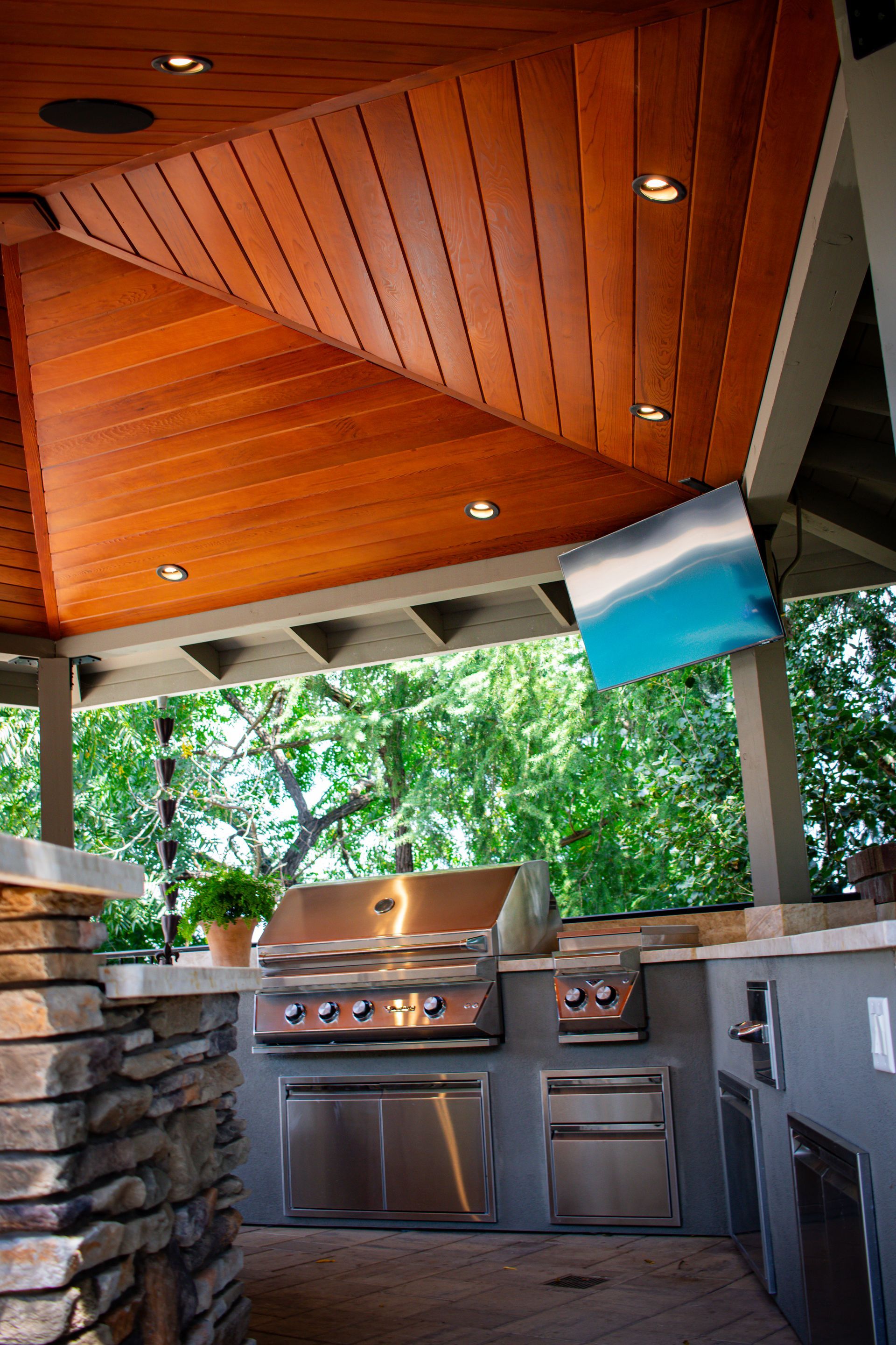Outdoor kitchen with stainless steel grill, TV, wood ceiling, and stone accents.