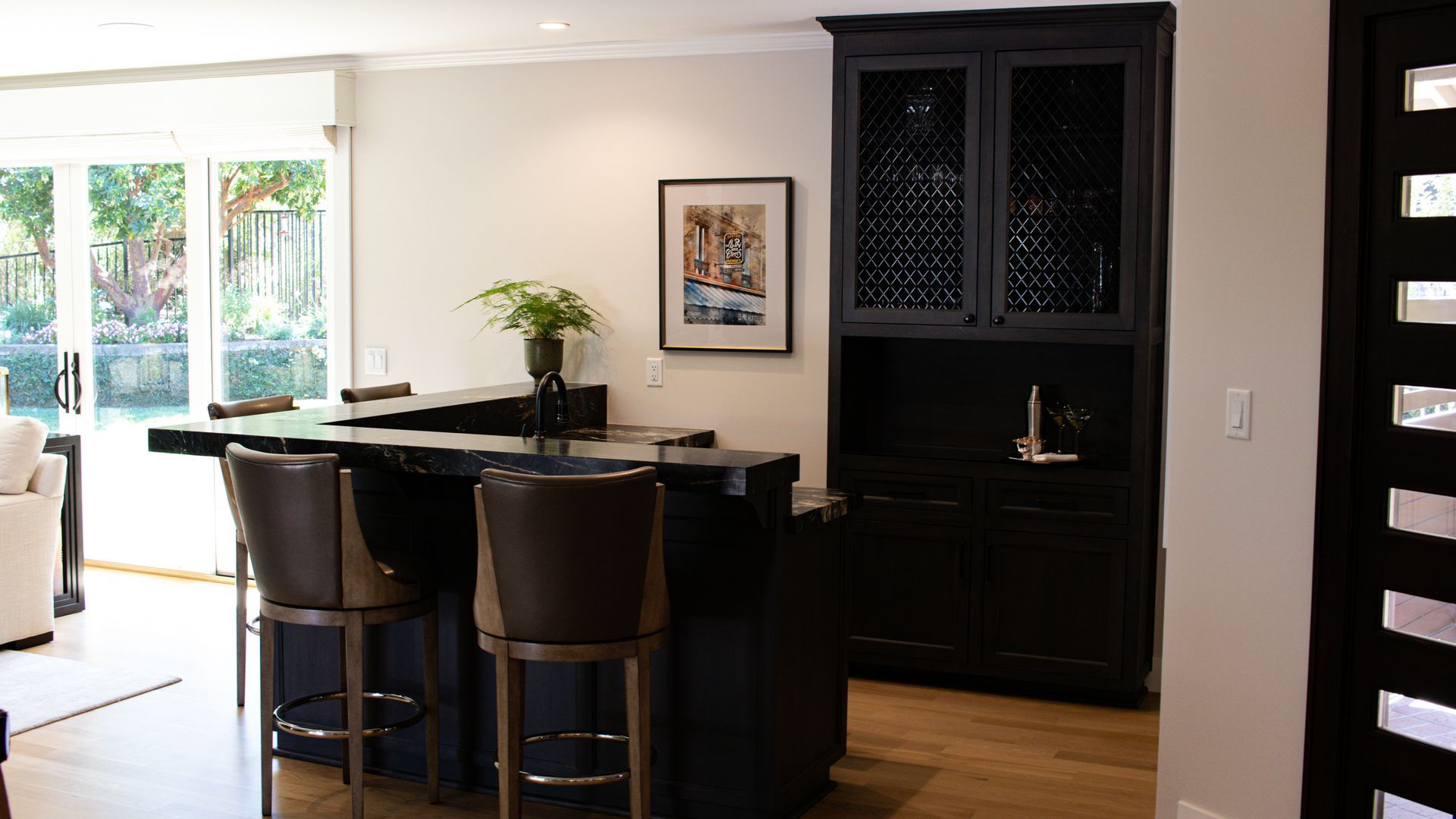 Bar area with black cabinets, stools, and a black countertop. A painting and plants are also visible.