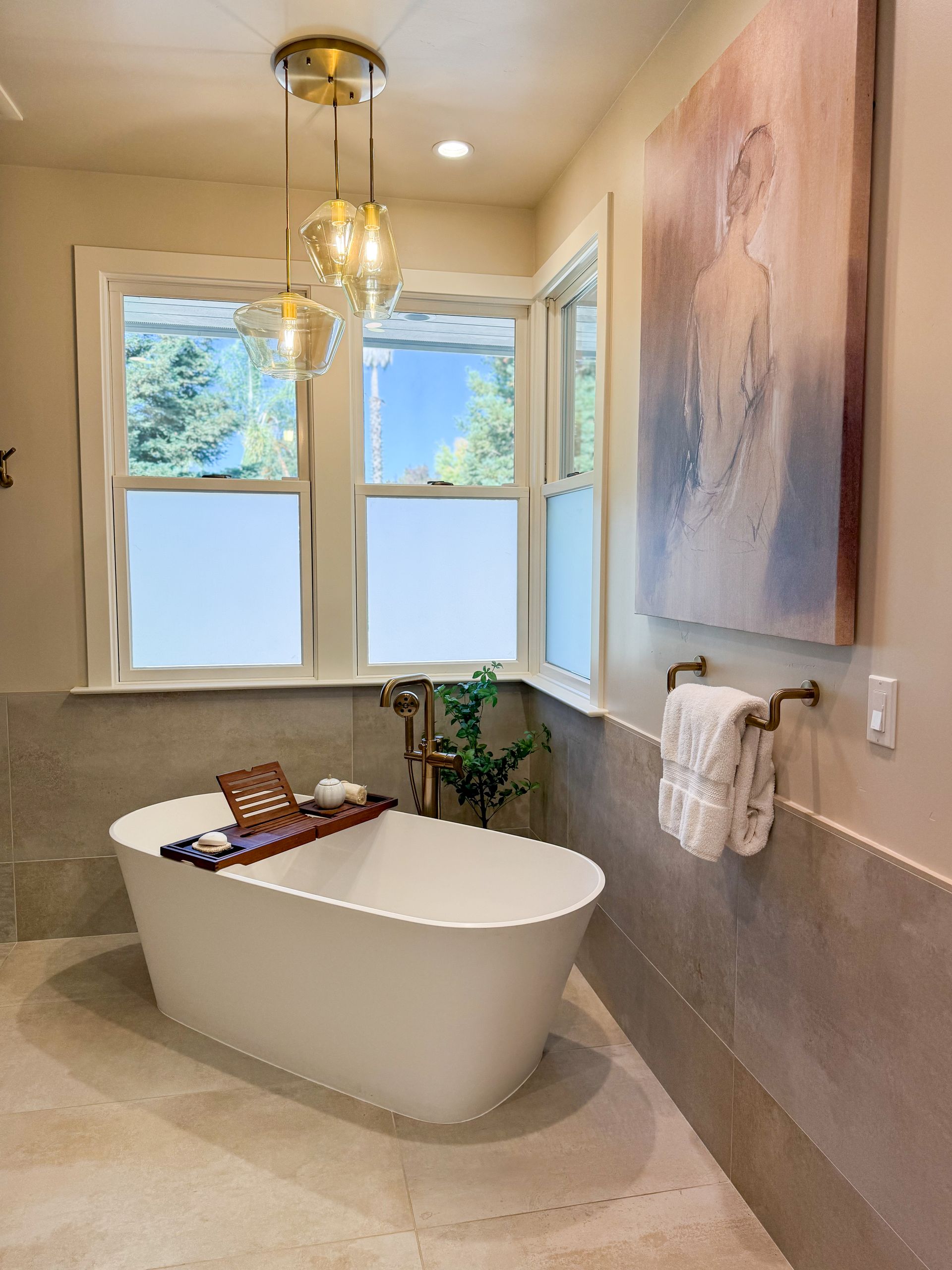 Bathroom with white tub, window, artwork, and gold light fixture.