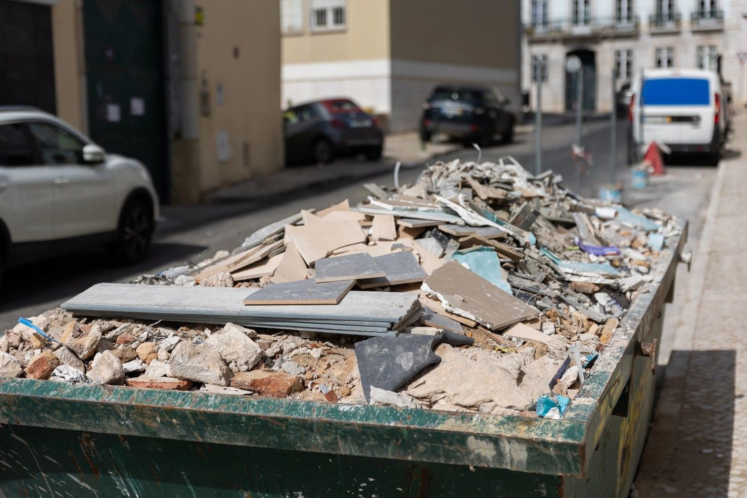 Un cassonetto pieno di detriti e macerie edili si trova su una strada cittadina vicino ad alcune auto parcheggiate.