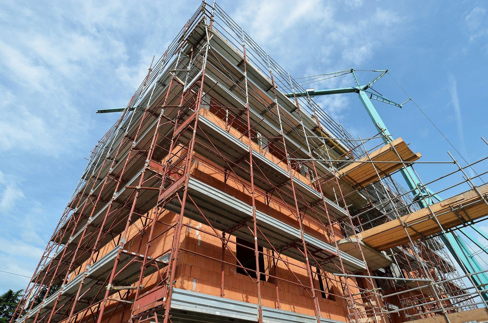 Un edificio in mattoni a più piani in costruzione, avvolto da impalcature metalliche, si staglia contro un cielo azzurro e nuvoloso.