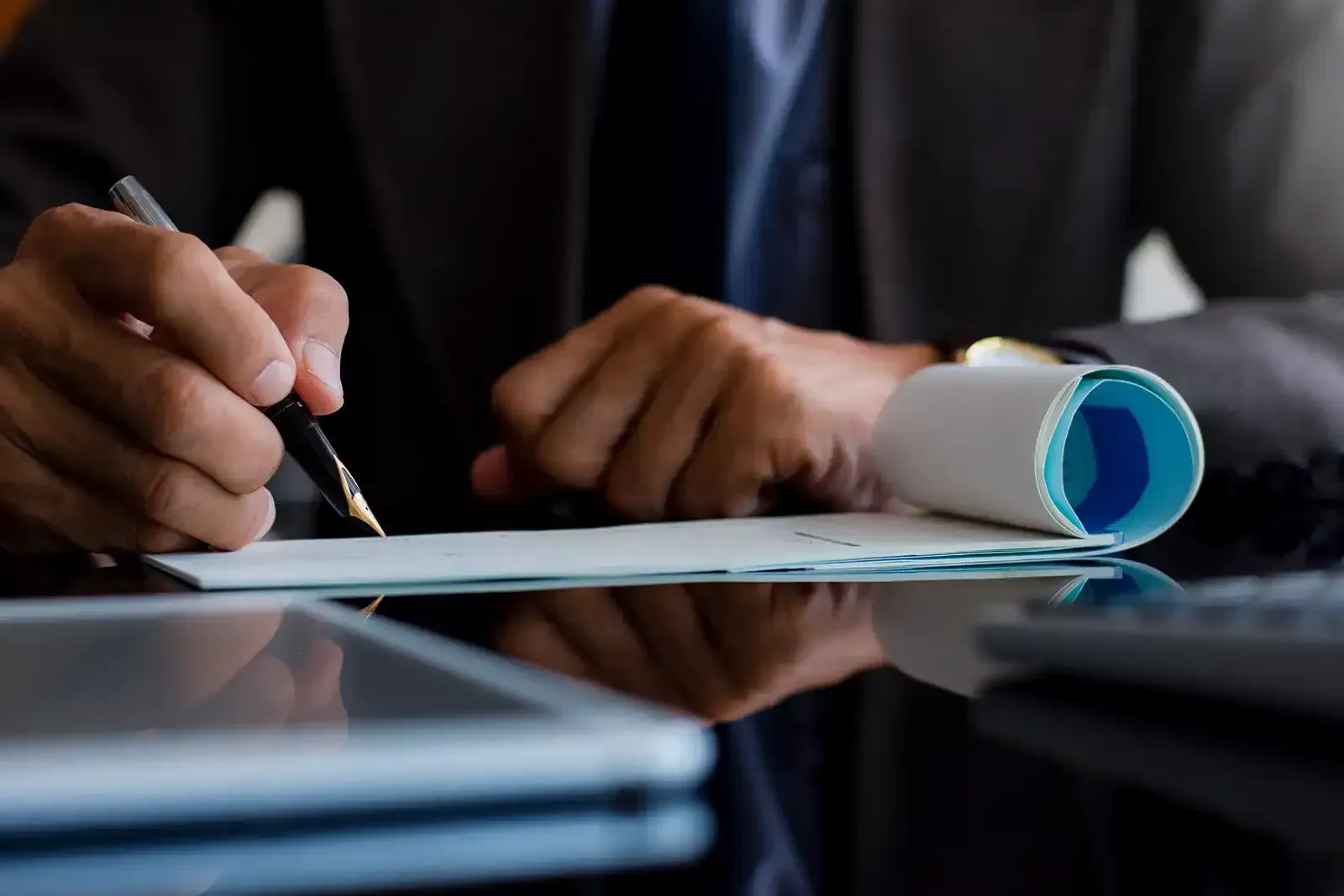 Person in suit writing on a check at a desk, near a tablet and calculator.