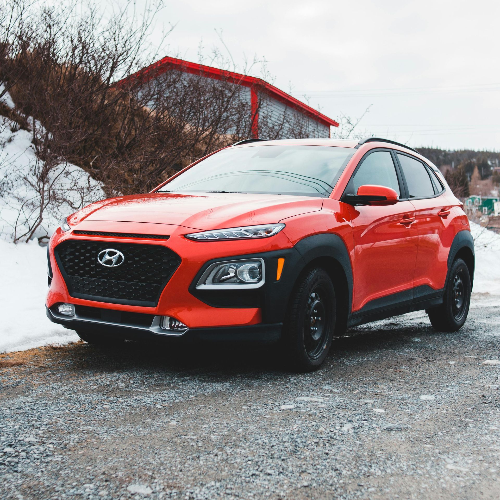 Red Hyundai Kona SUV parked on a gravel road in a winter setting.