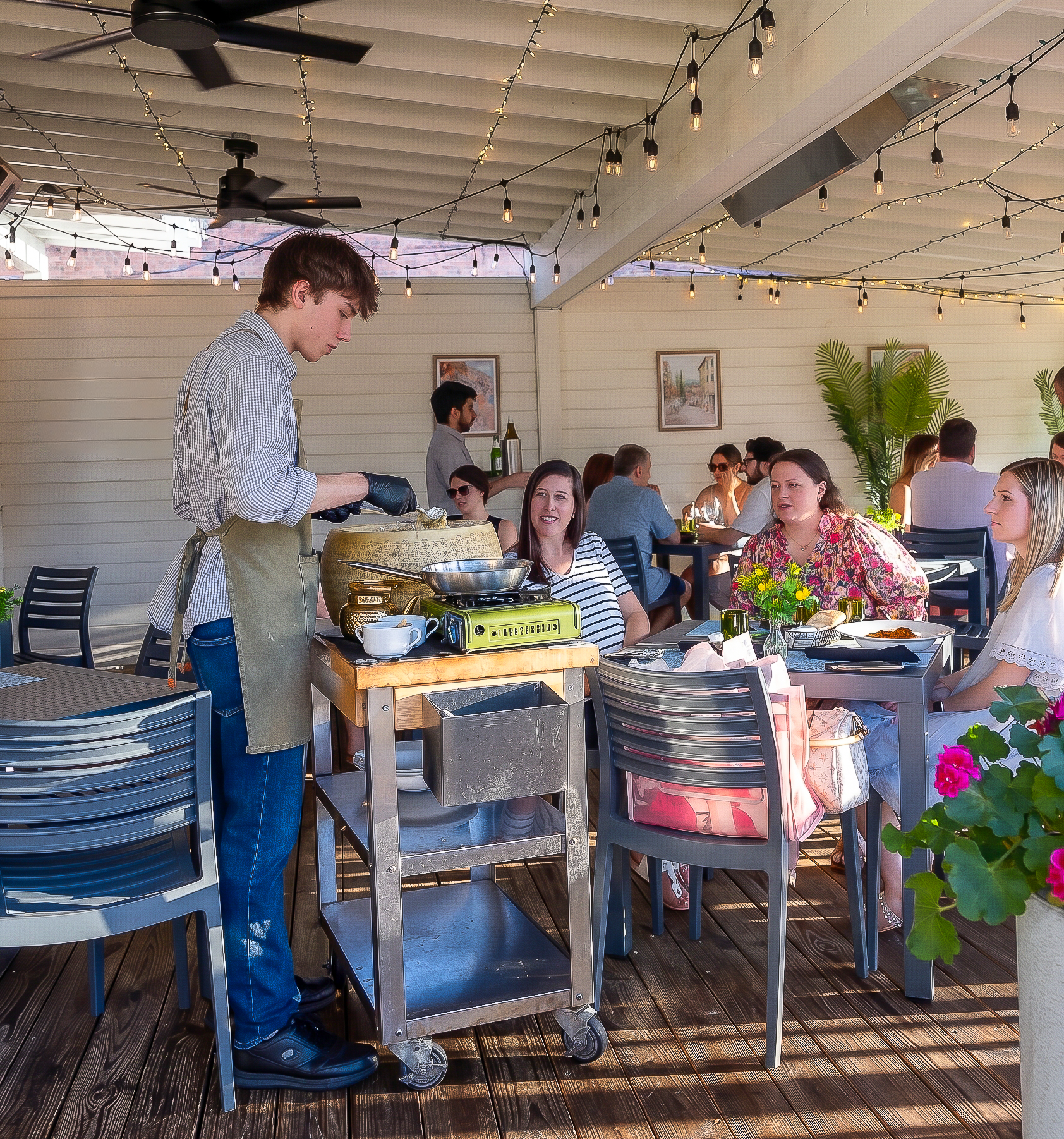 Server presenting food to diners at a sunlit outdoor cafe with string lights and potted flowers.