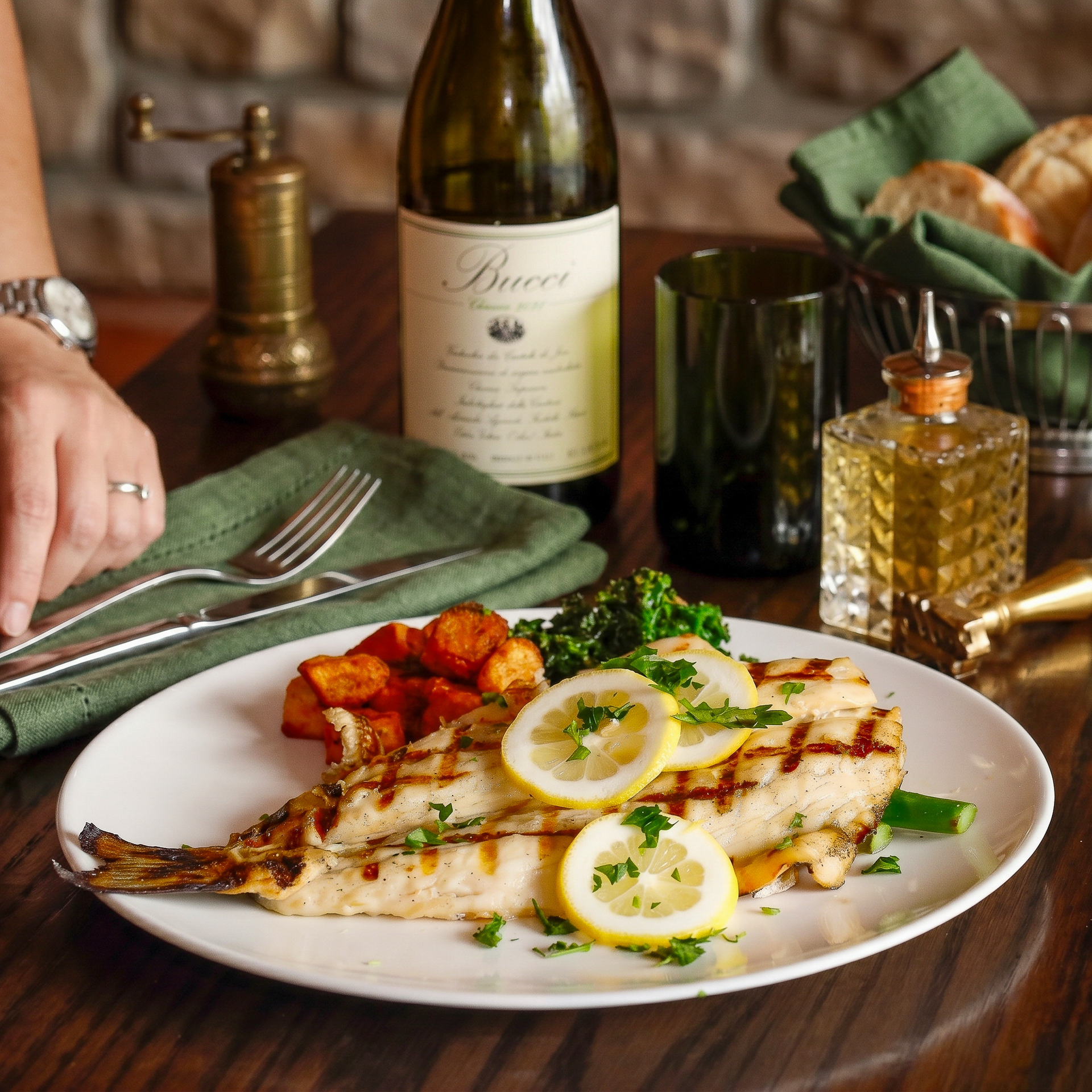 Grilled fish with lemon slices on a white plate, served on a table with wine and bread.