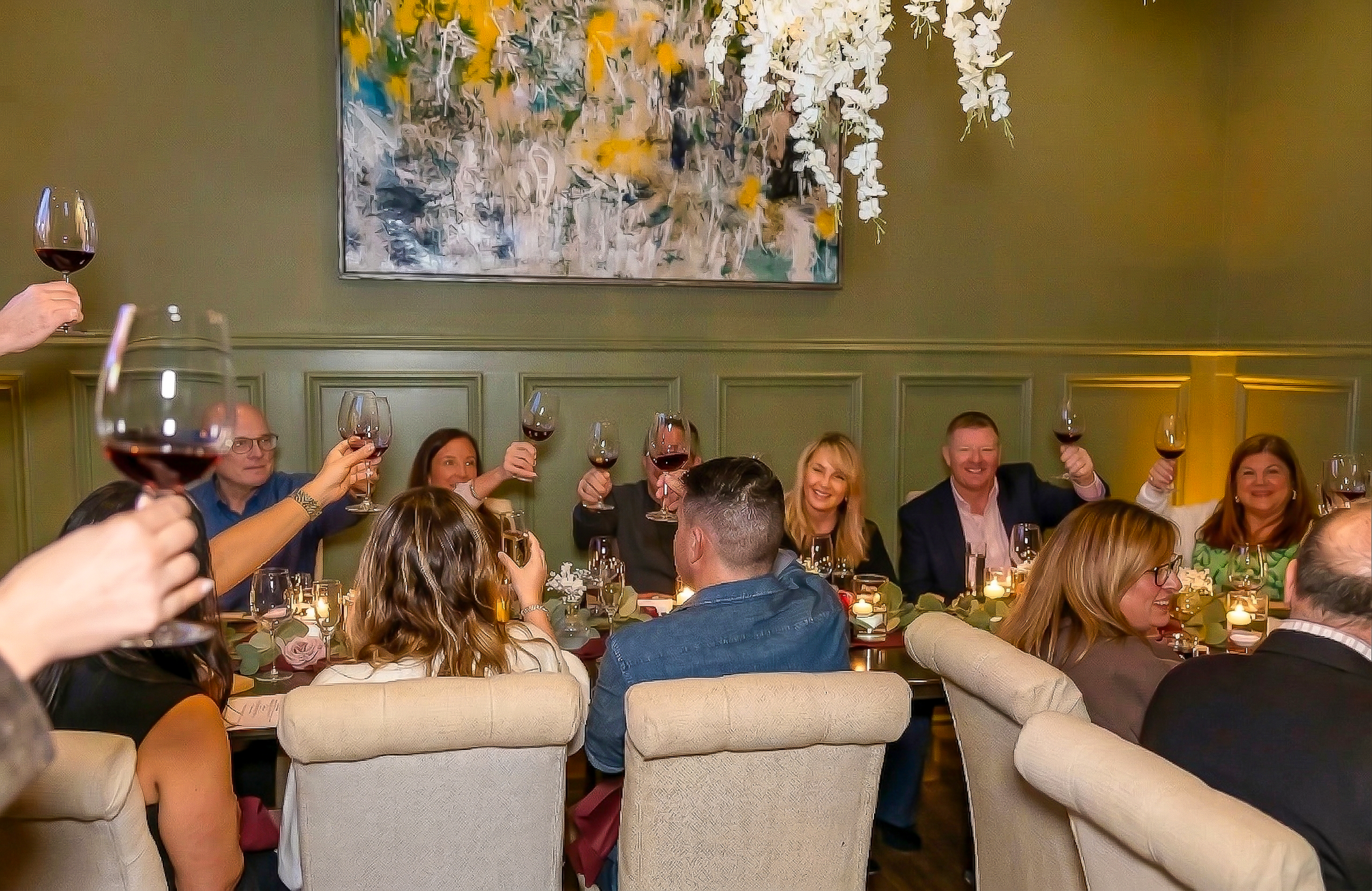 People seated in a lounge raise glasses during a social gathering, with warm lighting and wall art.