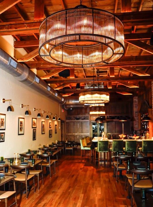 Warm-lit rustic restaurant interior with wooden tables, chairs, and large pendant lights.