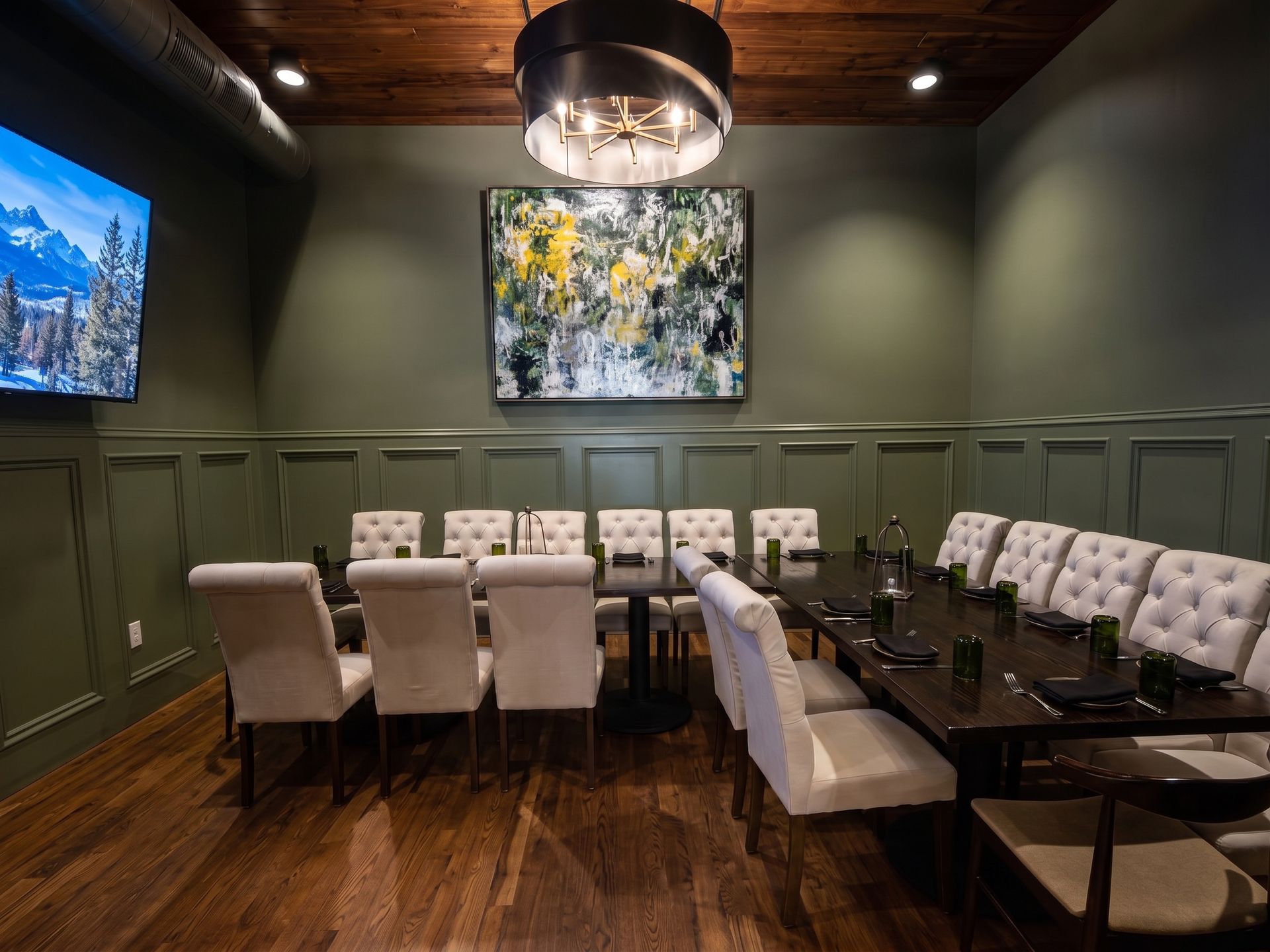 Modern dining room with a long table, white chairs, dark green walls, and framed art under a wood ceiling