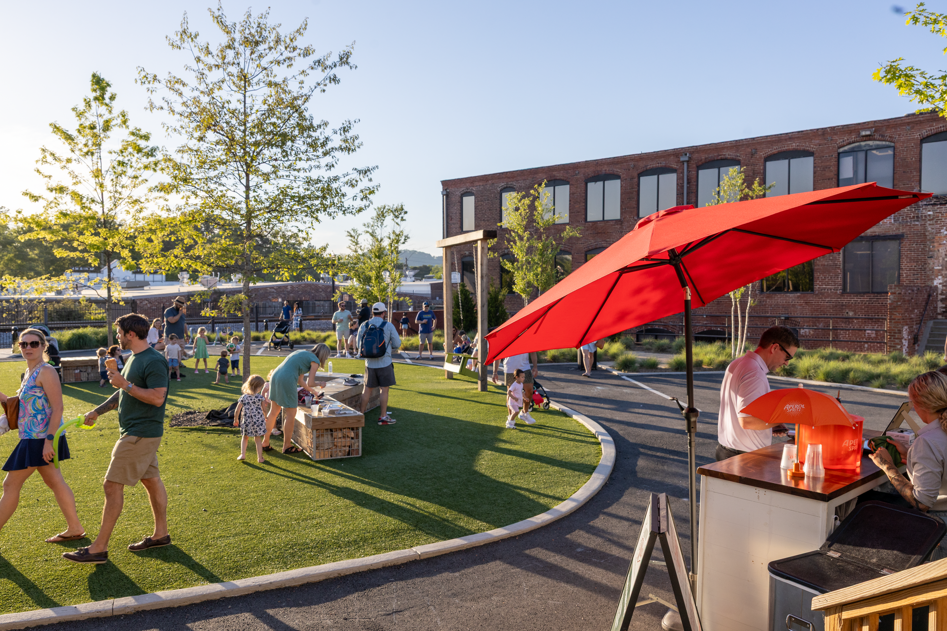 People relaxing outdoors on a sunny lawn beside a building, with red umbrellas and a grill nearby.
