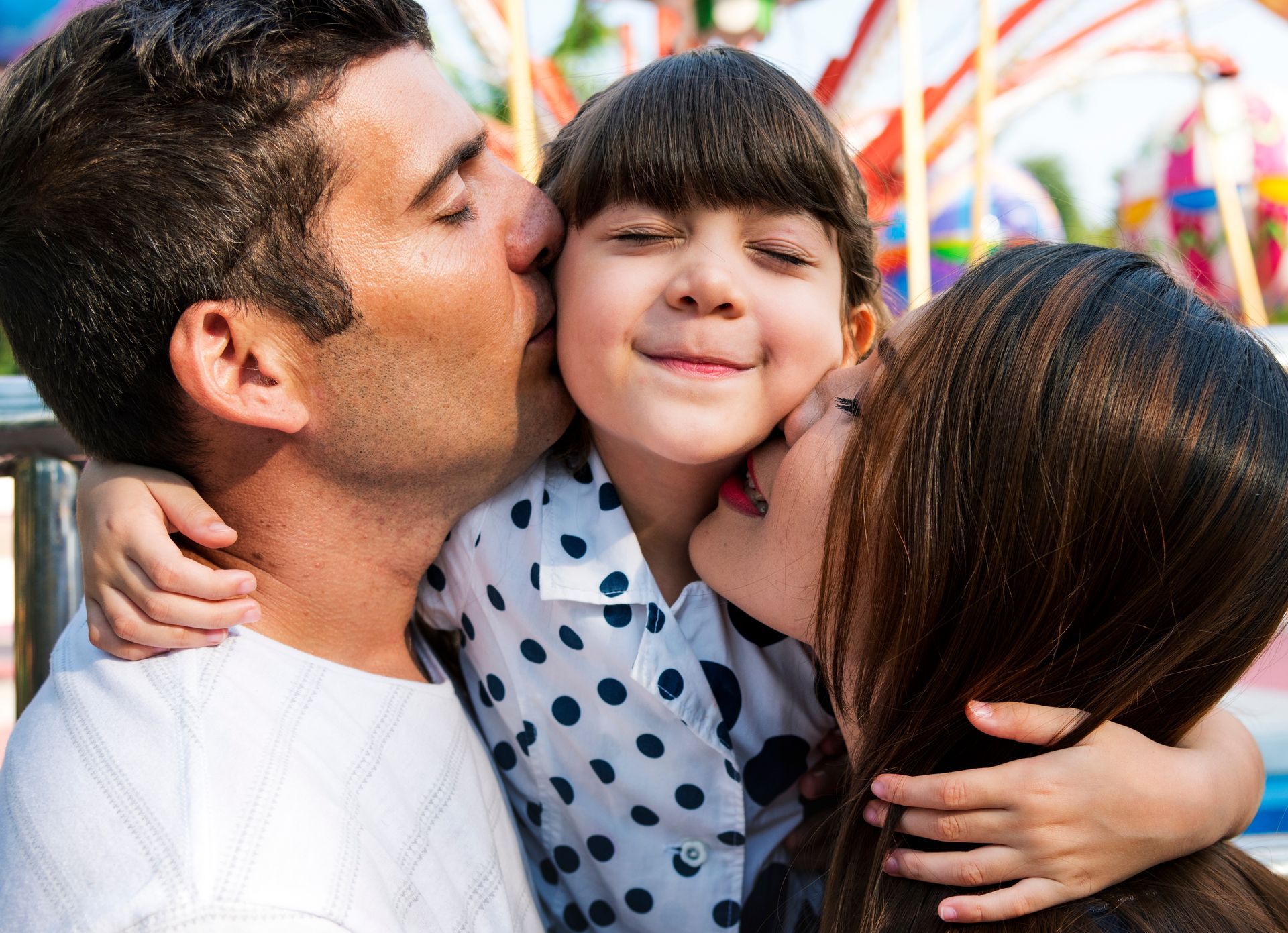 Two adults kissing a smiling child on the cheeks outdoors