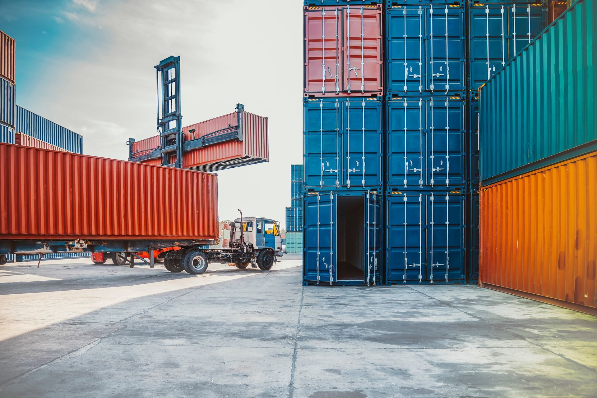 Container yard with stacked blue and orange shipping containers and a forklift near a red trailer