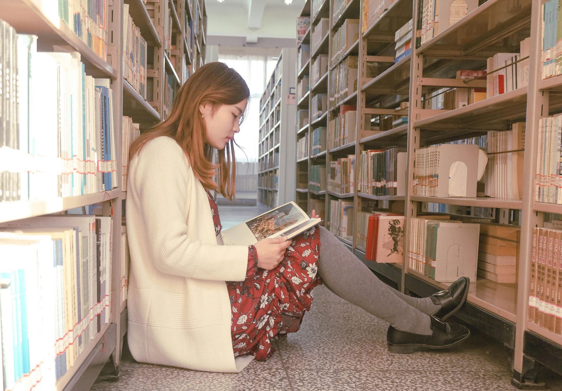A woman is sitting on the floor in a library reading a book.