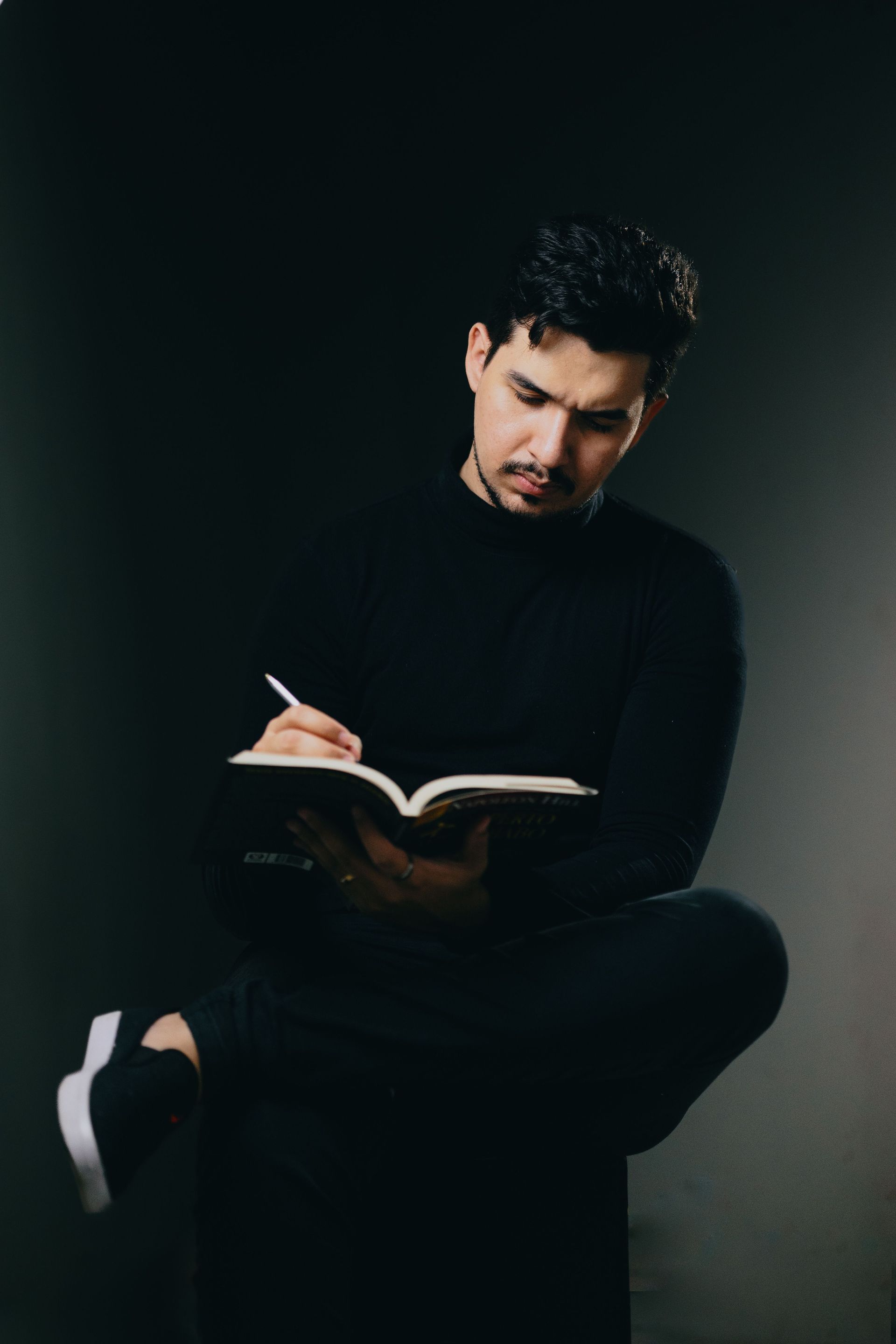 Person seated against a dark background, writing in an open notebook.