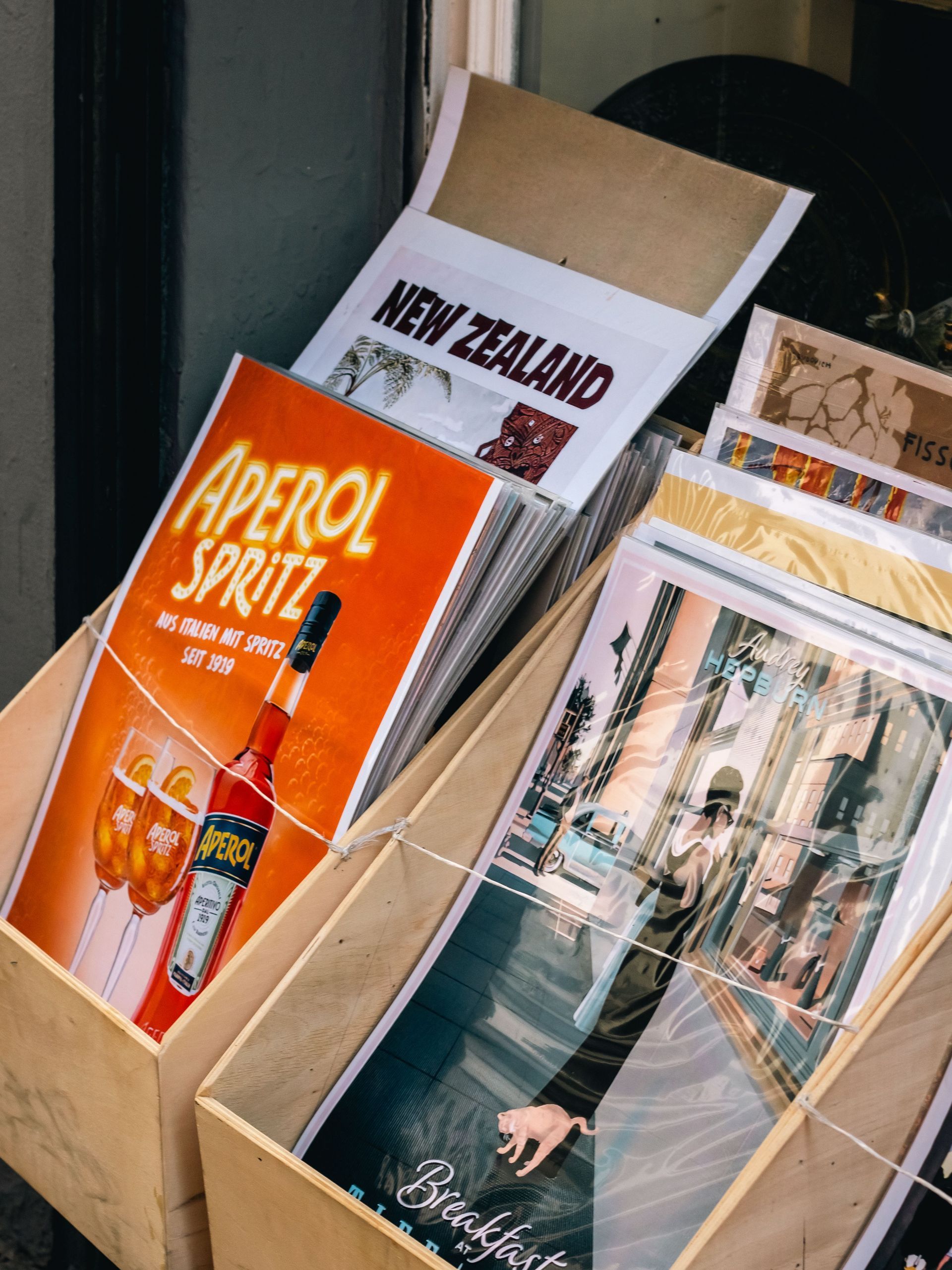 A cardboard box filled with colorful magazines and posters, including an Aperol Spritz ad and a New Zealand print.