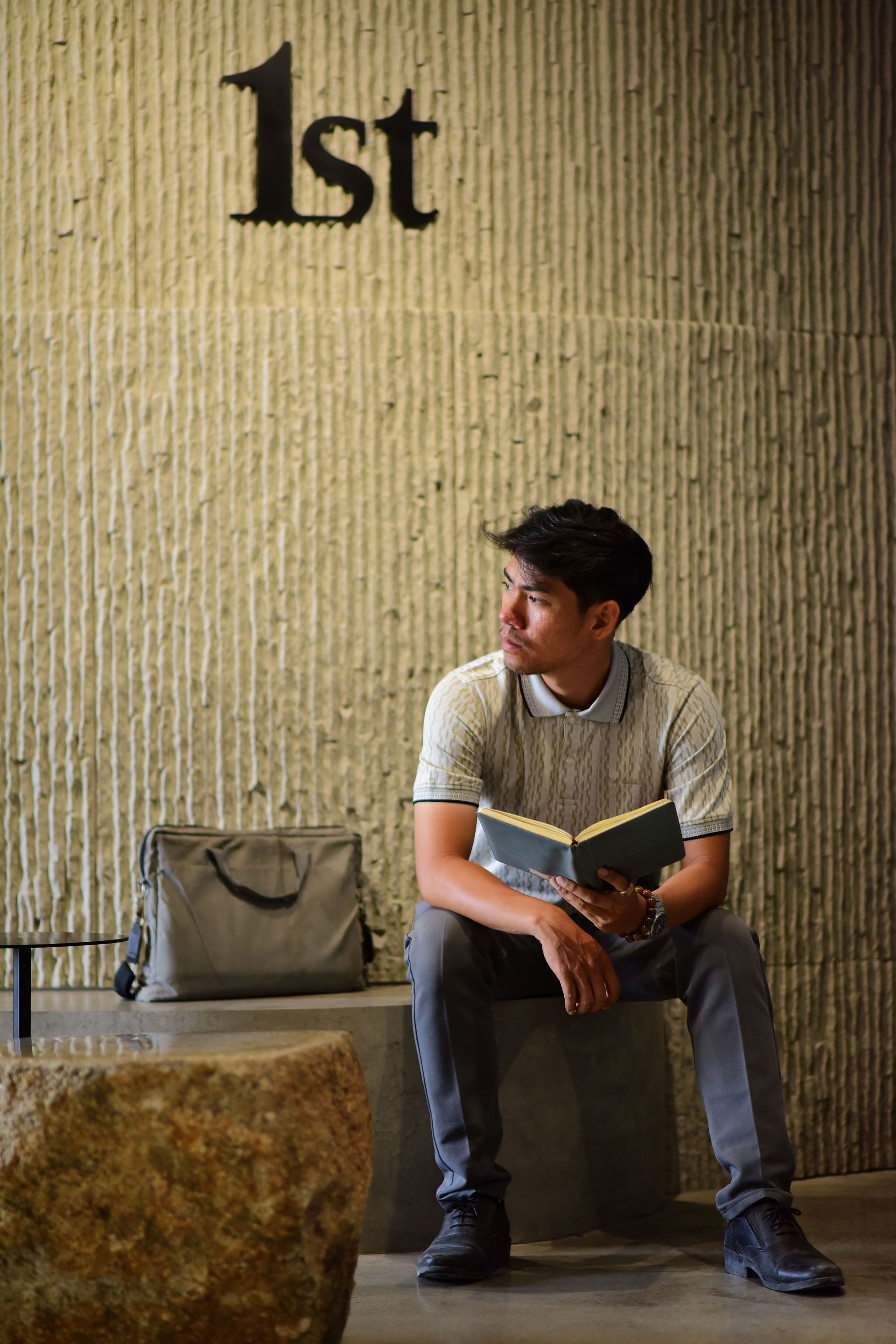 Person sitting on a bench reading a book beside a bag, with “1st” on the wall behind them.