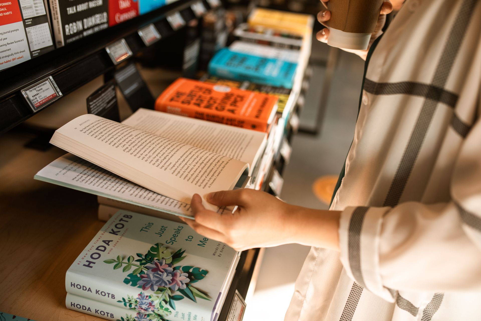 Person browsing books on a store shelf, holding an open book in a bookstore aisle