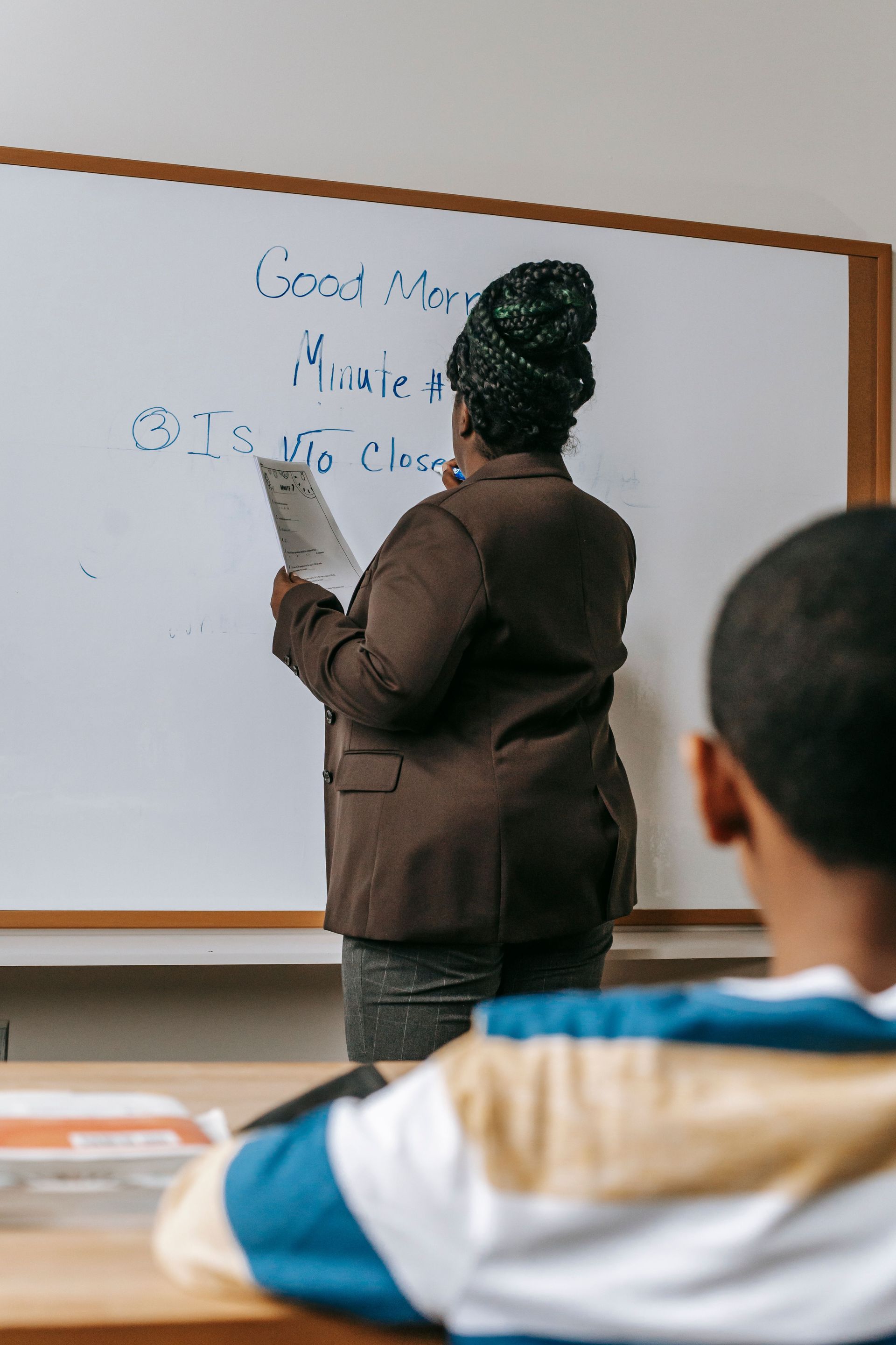 Teacher writing on a whiteboard while a student sits in the foreground.