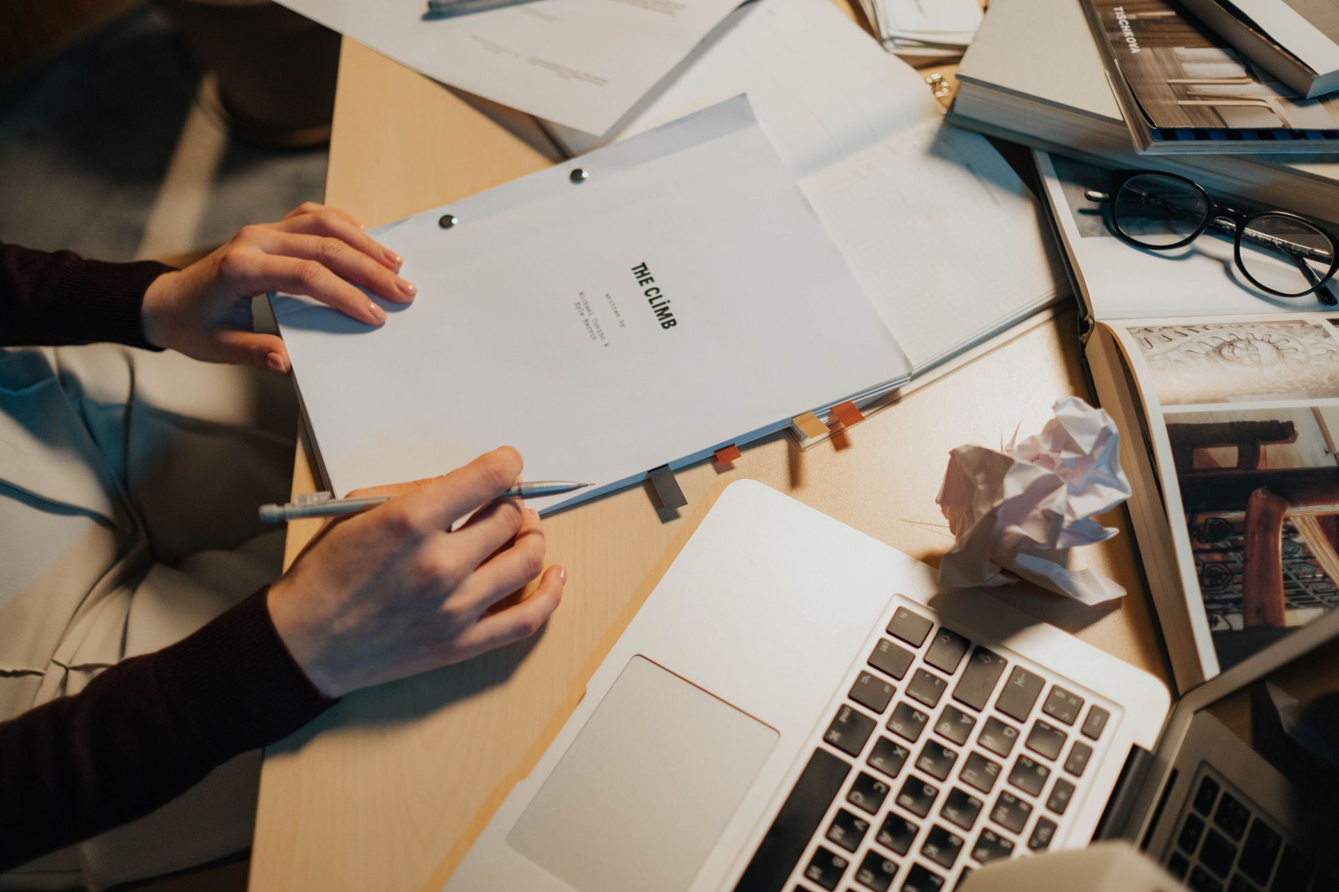 Hands sorting papers at a desk with a laptop, notebook, and crumpled paper.