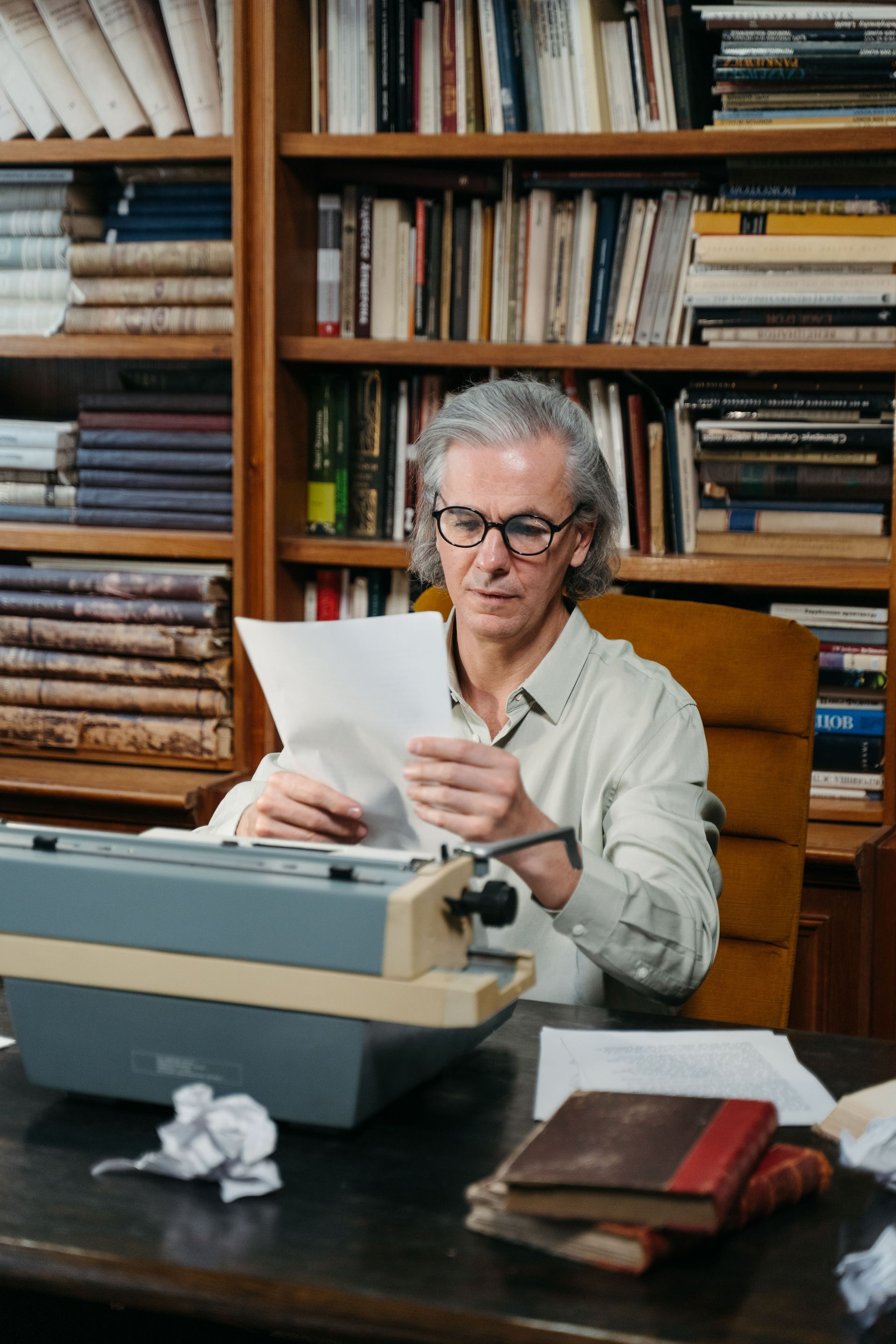 Person reading a paper at a desk beside a typewriter, with bookshelves in the background.