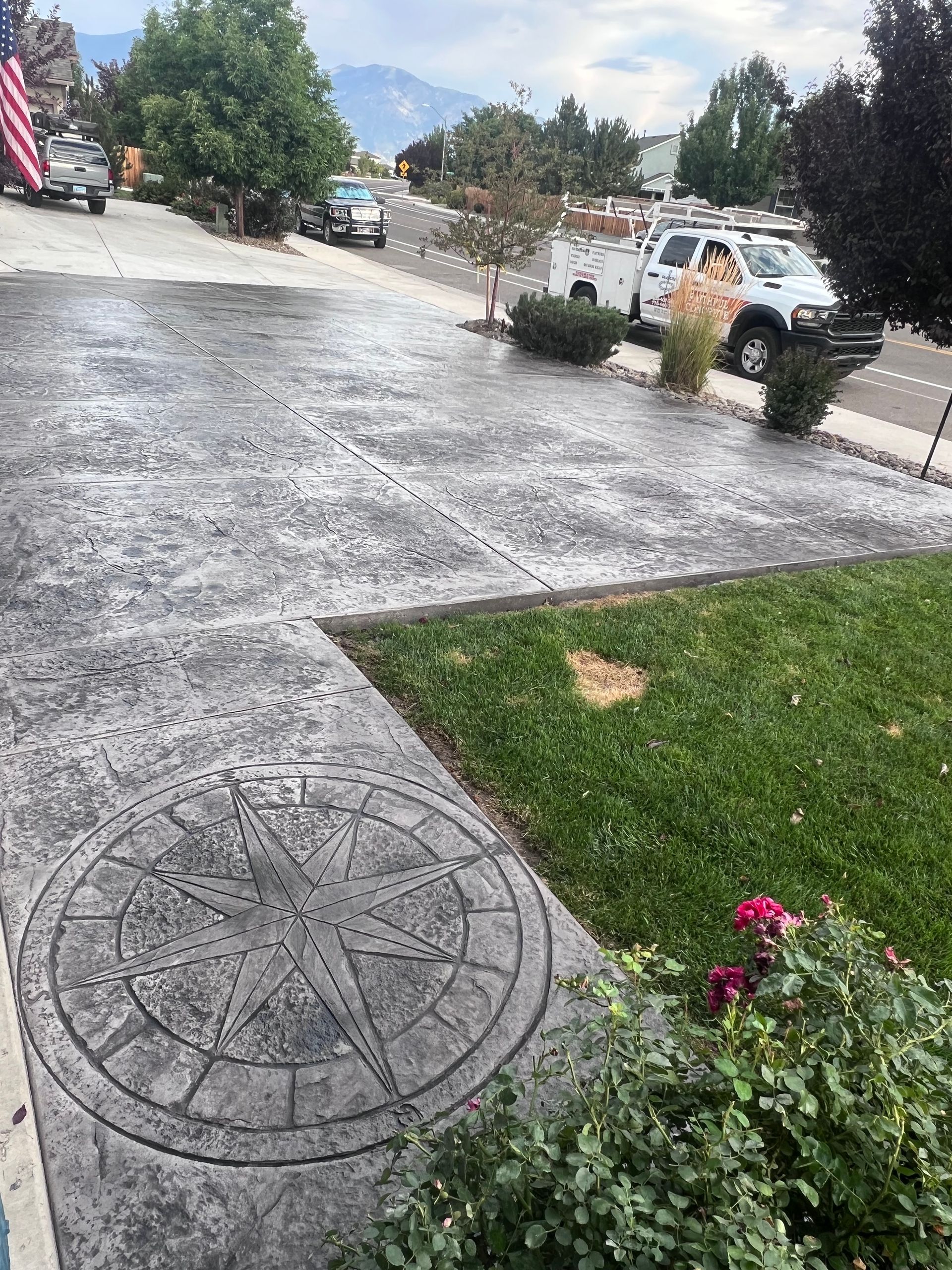 Decorative concrete driveway with compass medallion, bordered by lawn and flowers, with parked service vehicles nearby