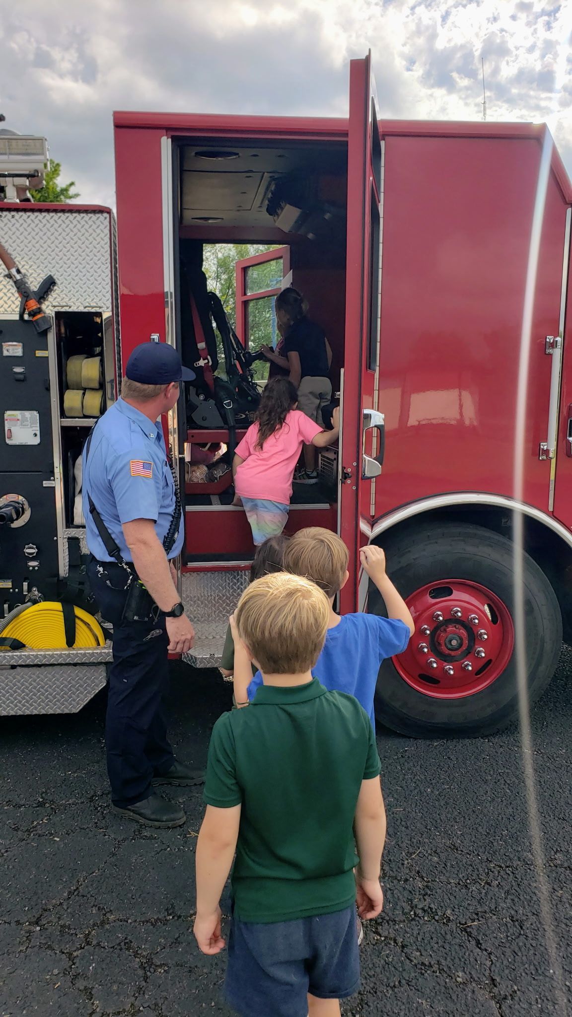 Firefighter with children exploring a red fire truck's interior; outside in daylight.