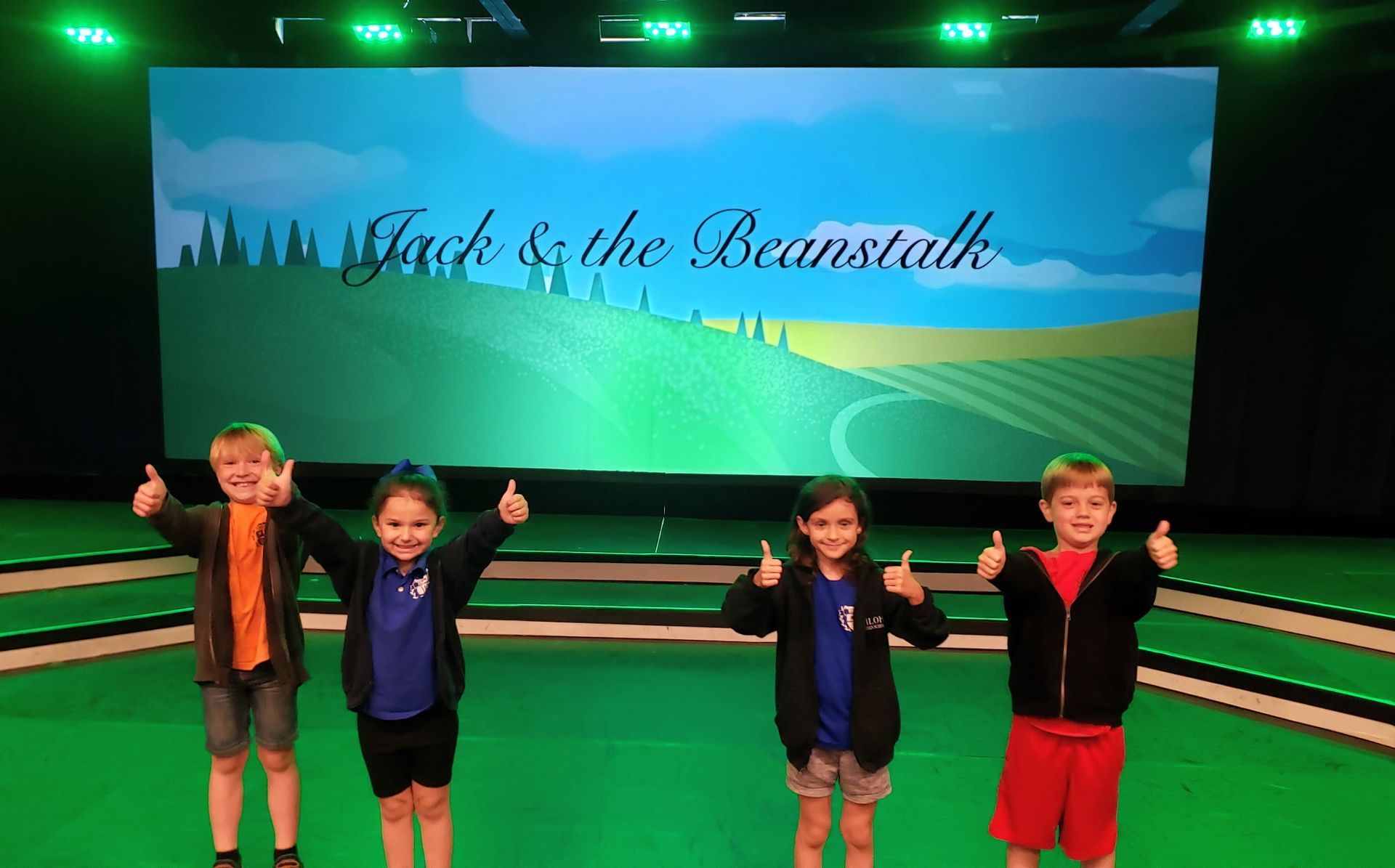 Four children on stage, thumbs up, in front of a backdrop that reads 