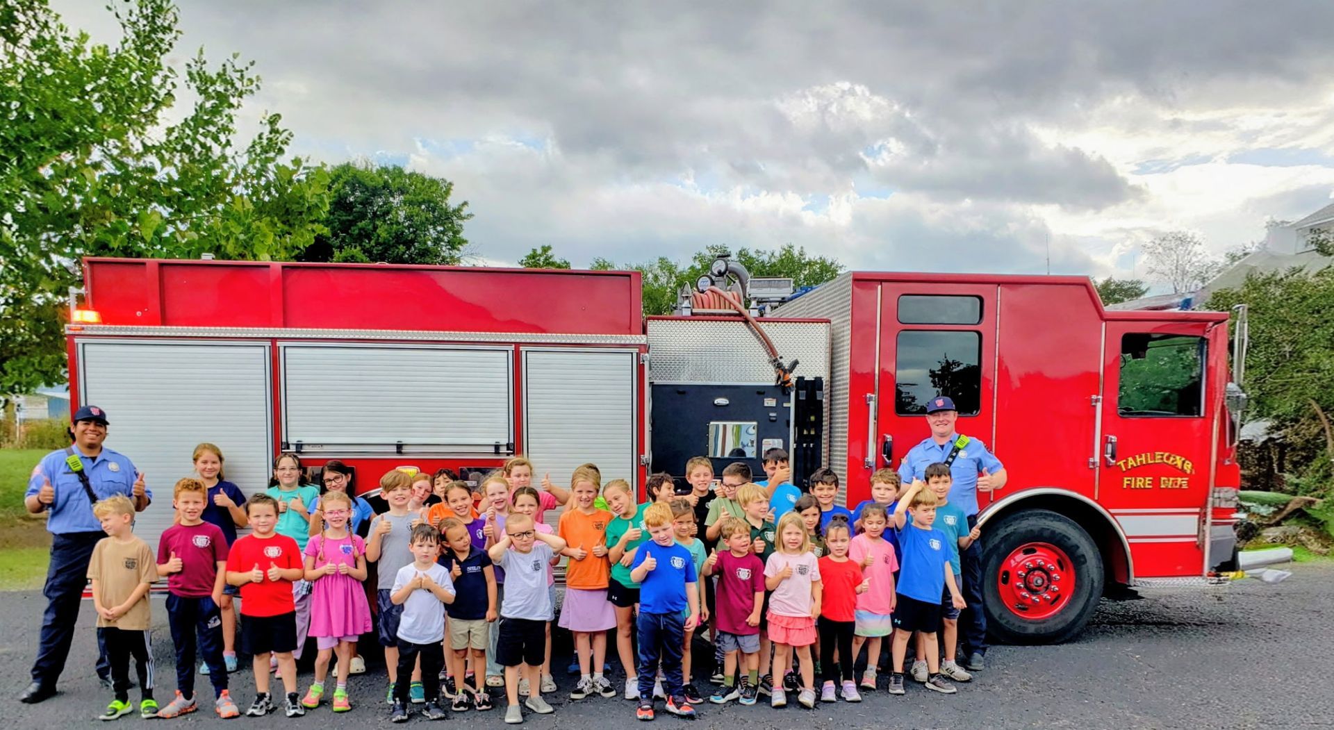 Children and firefighters pose in front of a red fire truck on a cloudy day.