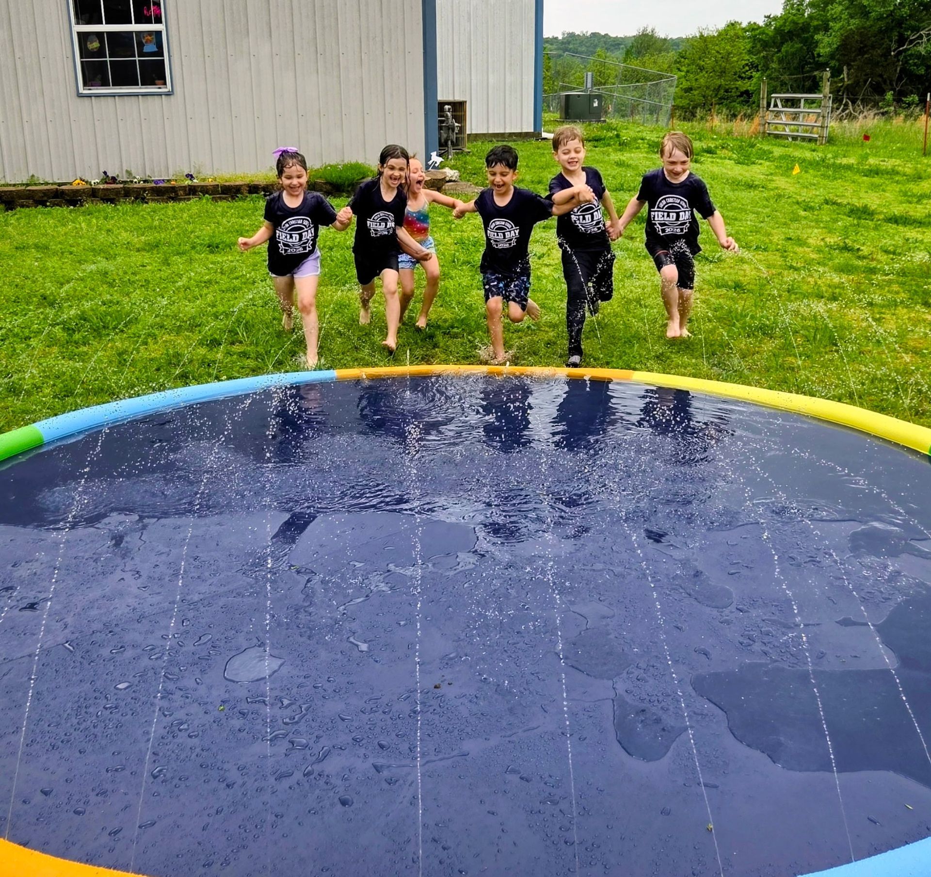 Five children running toward a water splash pad in a grassy yard after rain