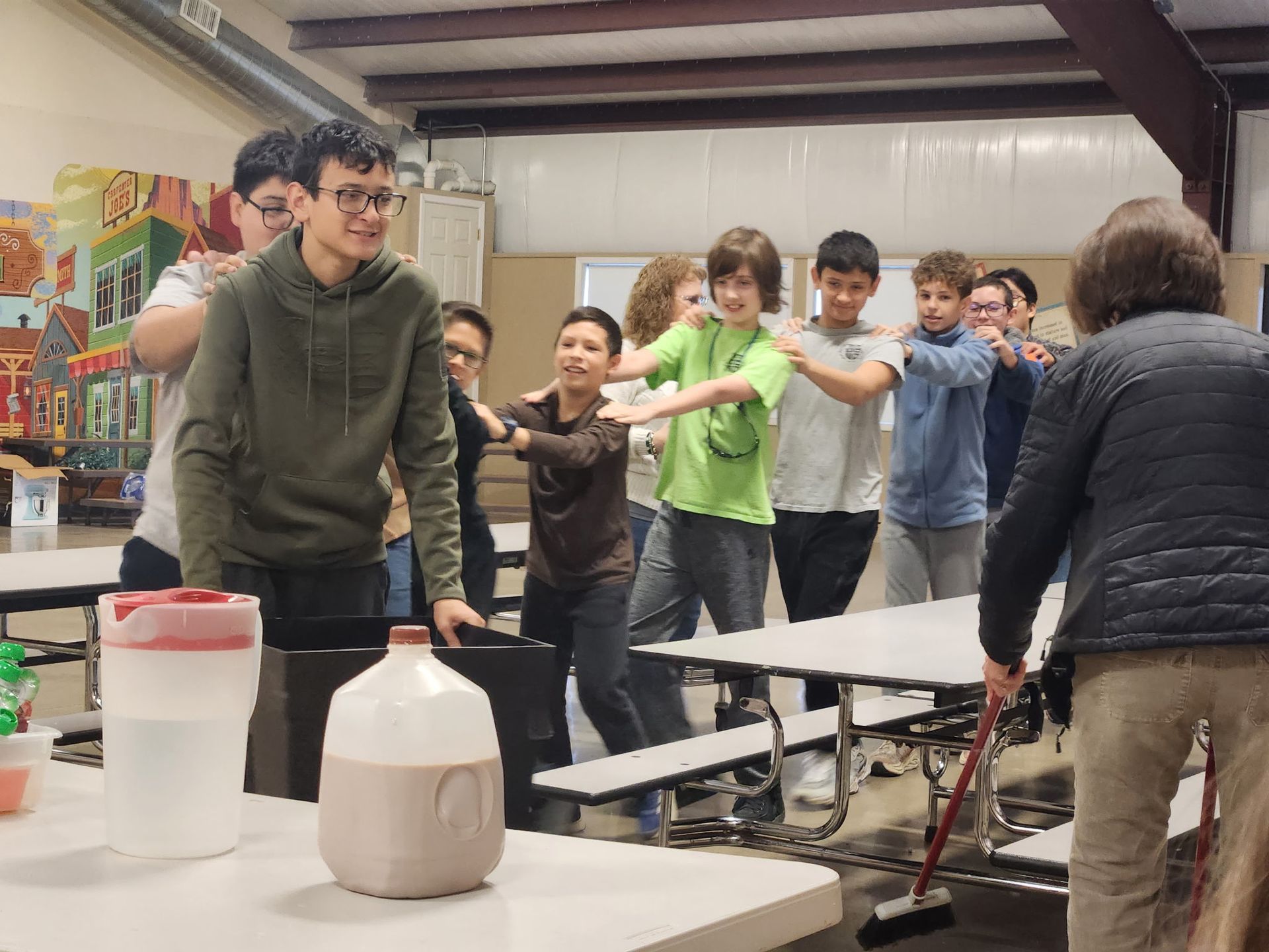 Group of people in a cafeteria, playing a game, some with arms out, other looking on.