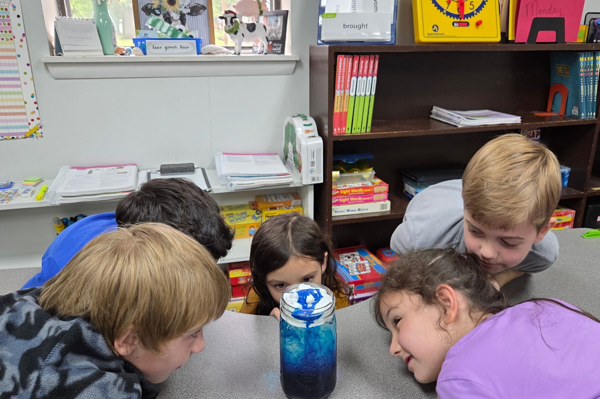 Children gathered around a table in a classroom, peering at a blue bottle center object.
