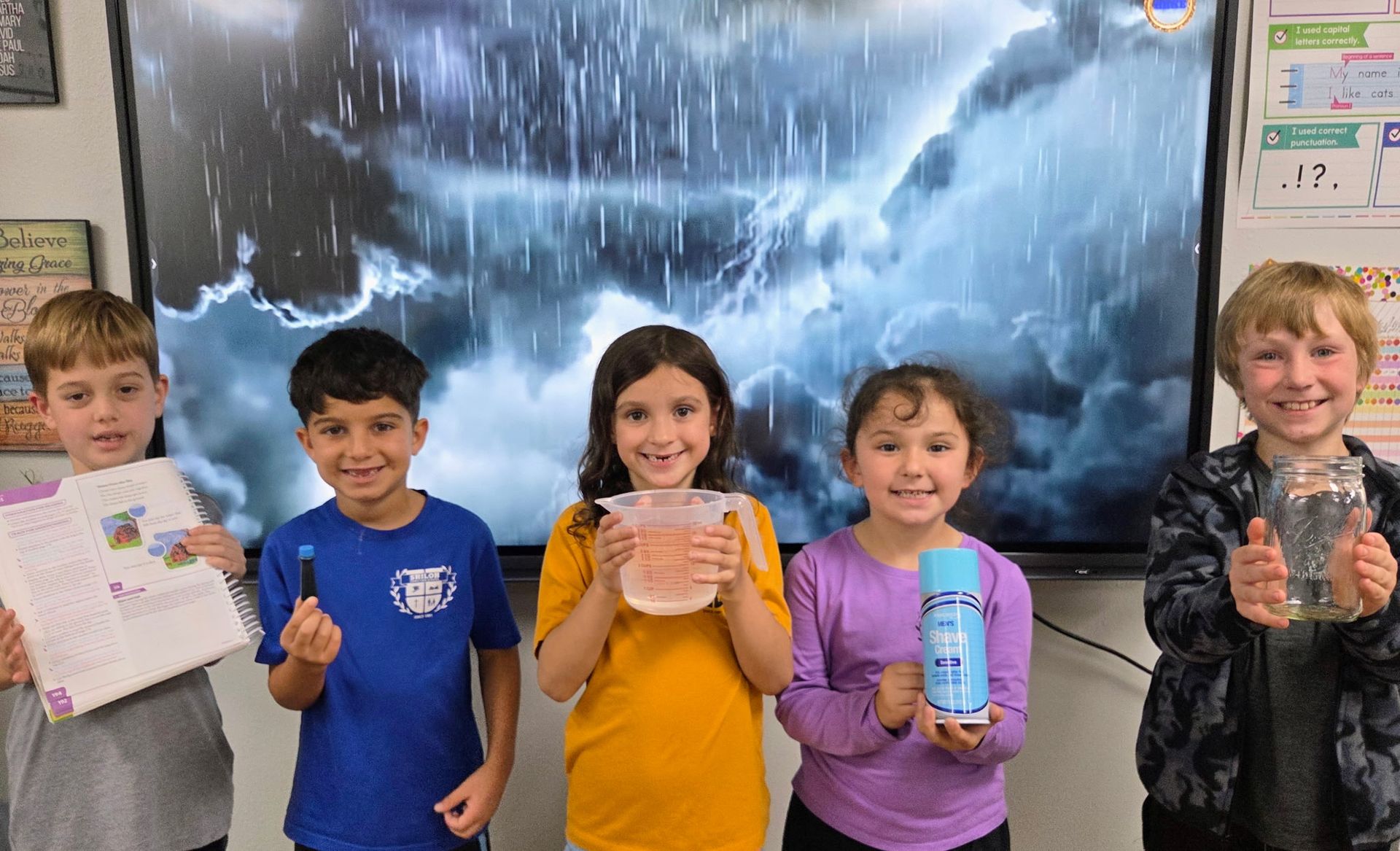 Five children stand in a classroom holding science projects in front of a weather poster.