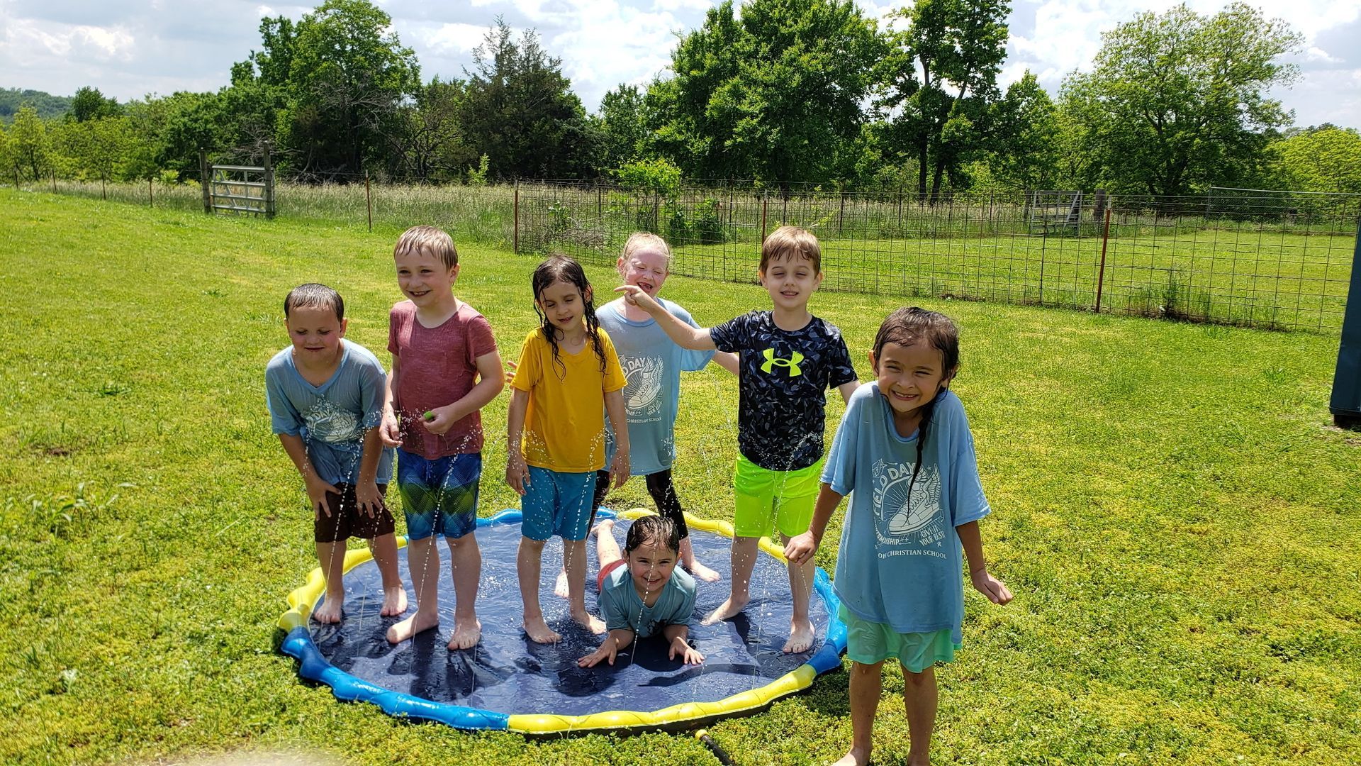 Children playing on a blue water pad in a grassy field on a sunny day.