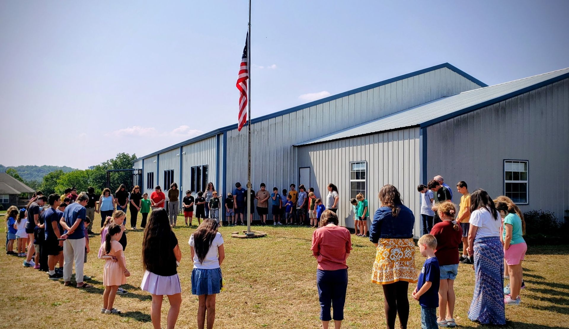 Students stand at attention in front of a school, flag at half-mast.