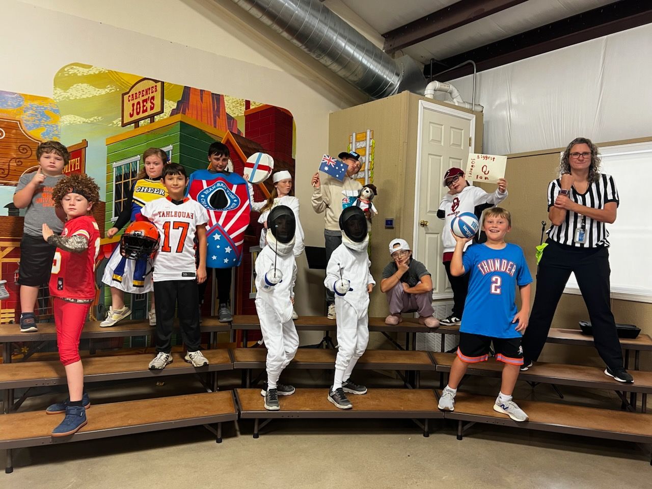 Children in sports costumes standing on a stage, with an astronaut and referee beside colorful boxes