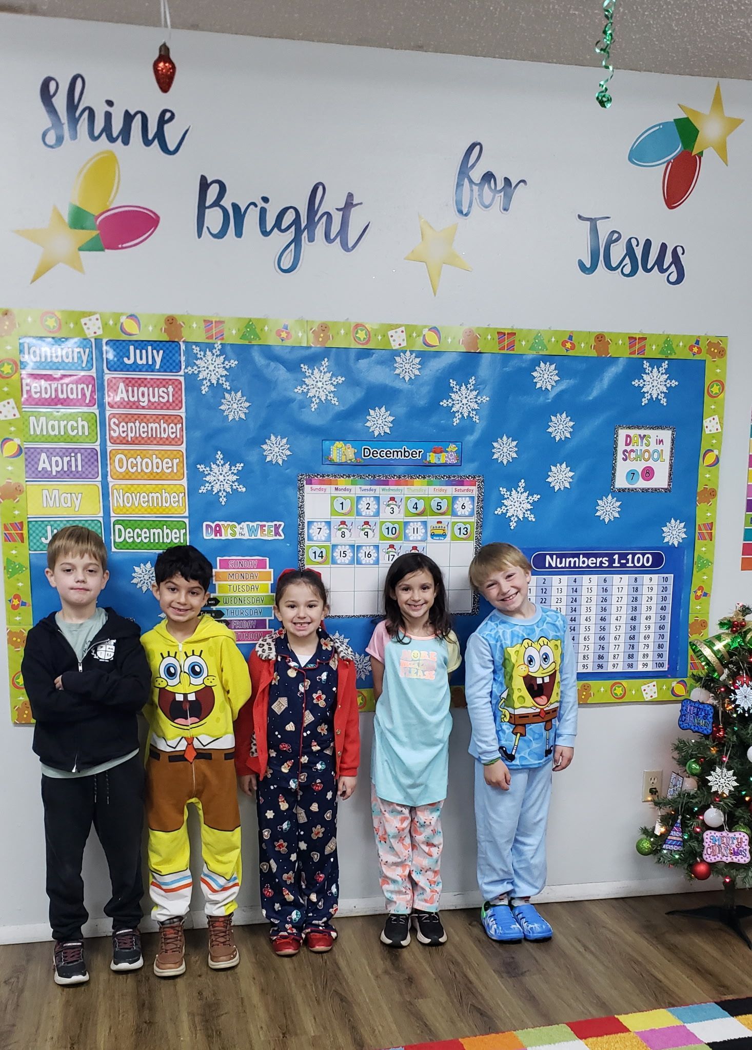 Children standing in a festive classroom with a winter bulletin board and a small decorated tree